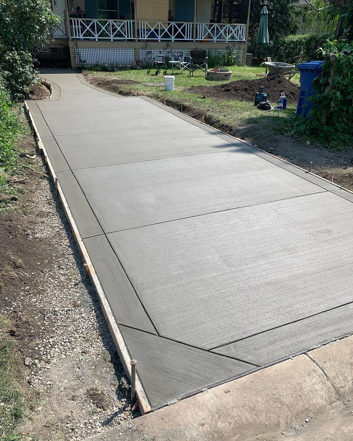Newly poured concrete sidewalk in a backyard with construction tools and supplies nearby, leading to a house with a porch and outdoor chairs.