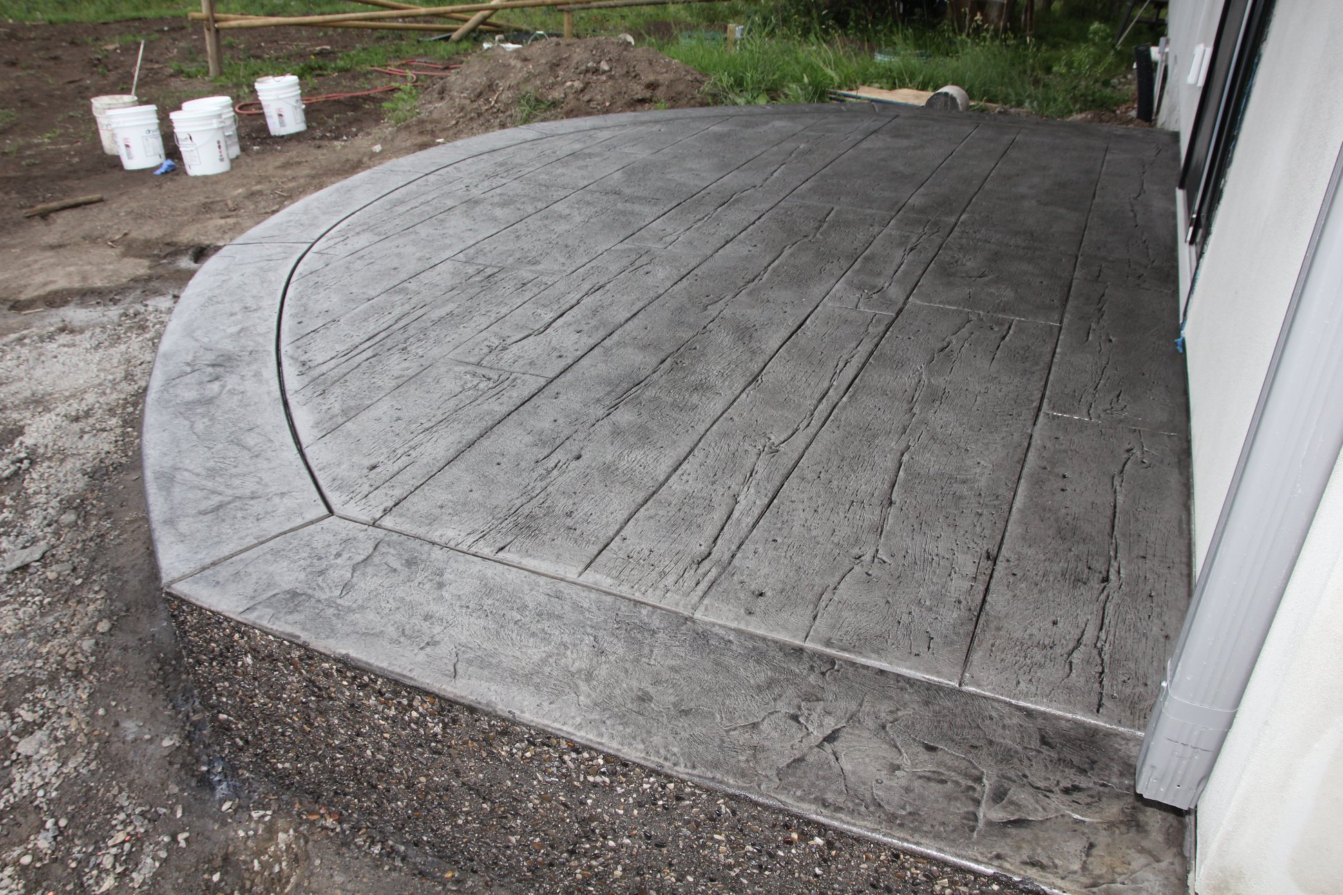 A newly constructed concrete patio with a textured, wood-like surface in front of a building, surrounded by construction materials and dirt.