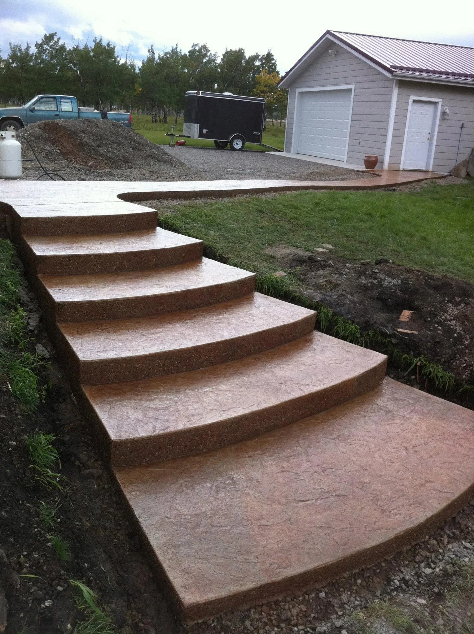 Newly installed outdoor concrete steps leading from yard to driveway, with a garage, lawn, and parked trailer in background.