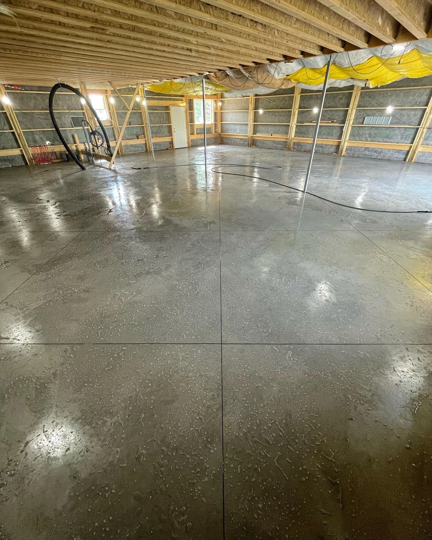 Interior of a house under construction with a newly poured concrete floor, visible insulation, wooden framing, electrical wiring, and some construction materials.