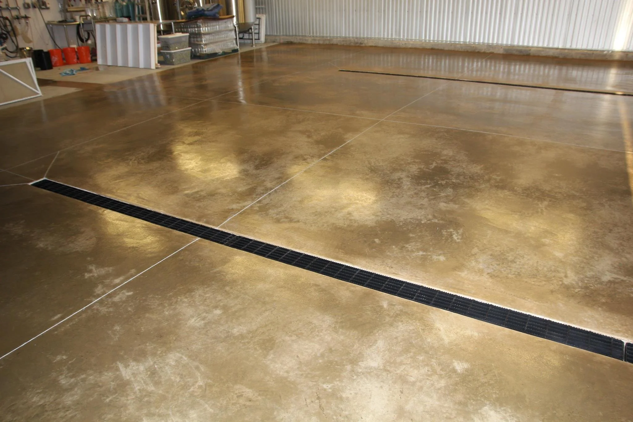Concrete floor with a black drainage grate in a workshop or warehouse with various tools and supplies in the background.