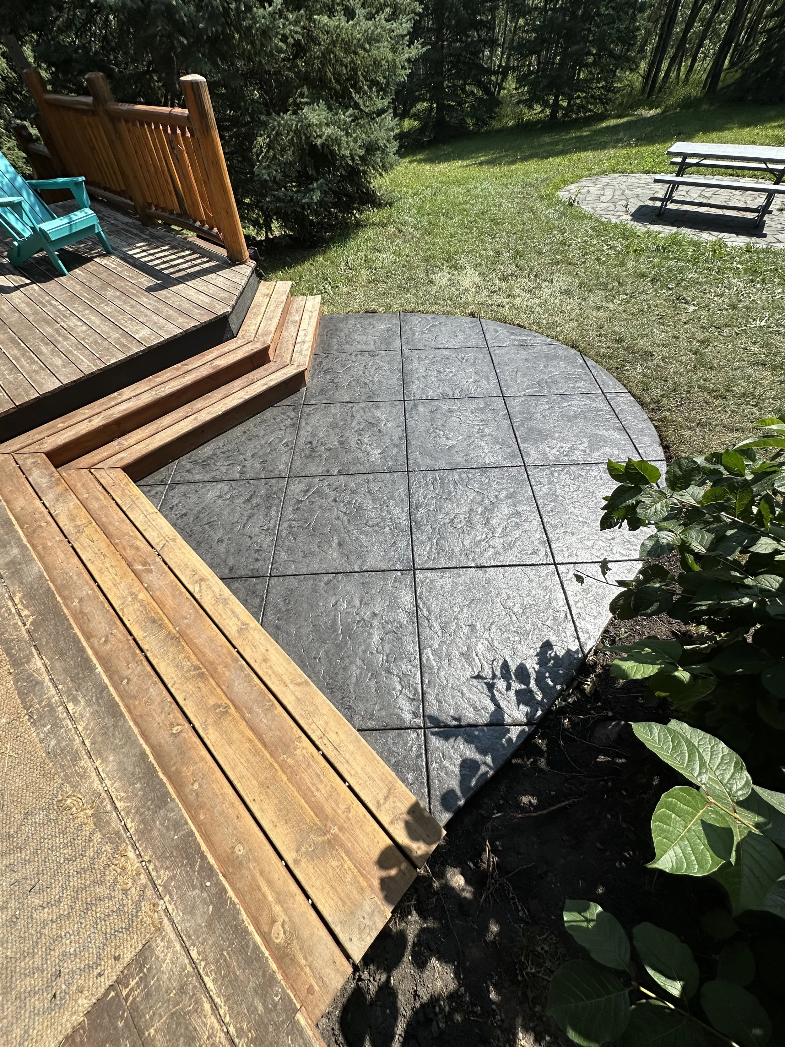 View of a backyard patio with a wooden deck, steps, and a stamped concrete area, surrounded by grass, bushes, and trees, with benches on a stone-paved section.