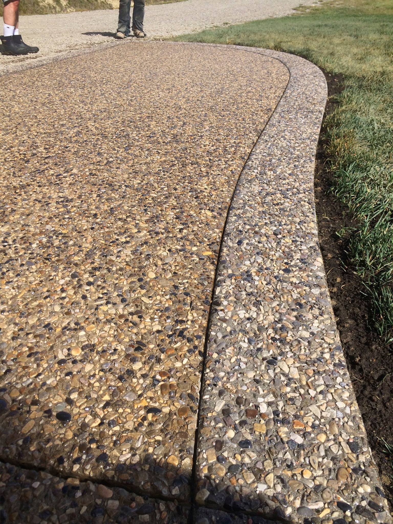 Close-up of a curved concrete sidewalk with a pebble surface, bordered by a strip of dirt and grass on the right side. Two people wearing boots and sneakers are partially visible at the top of the image.