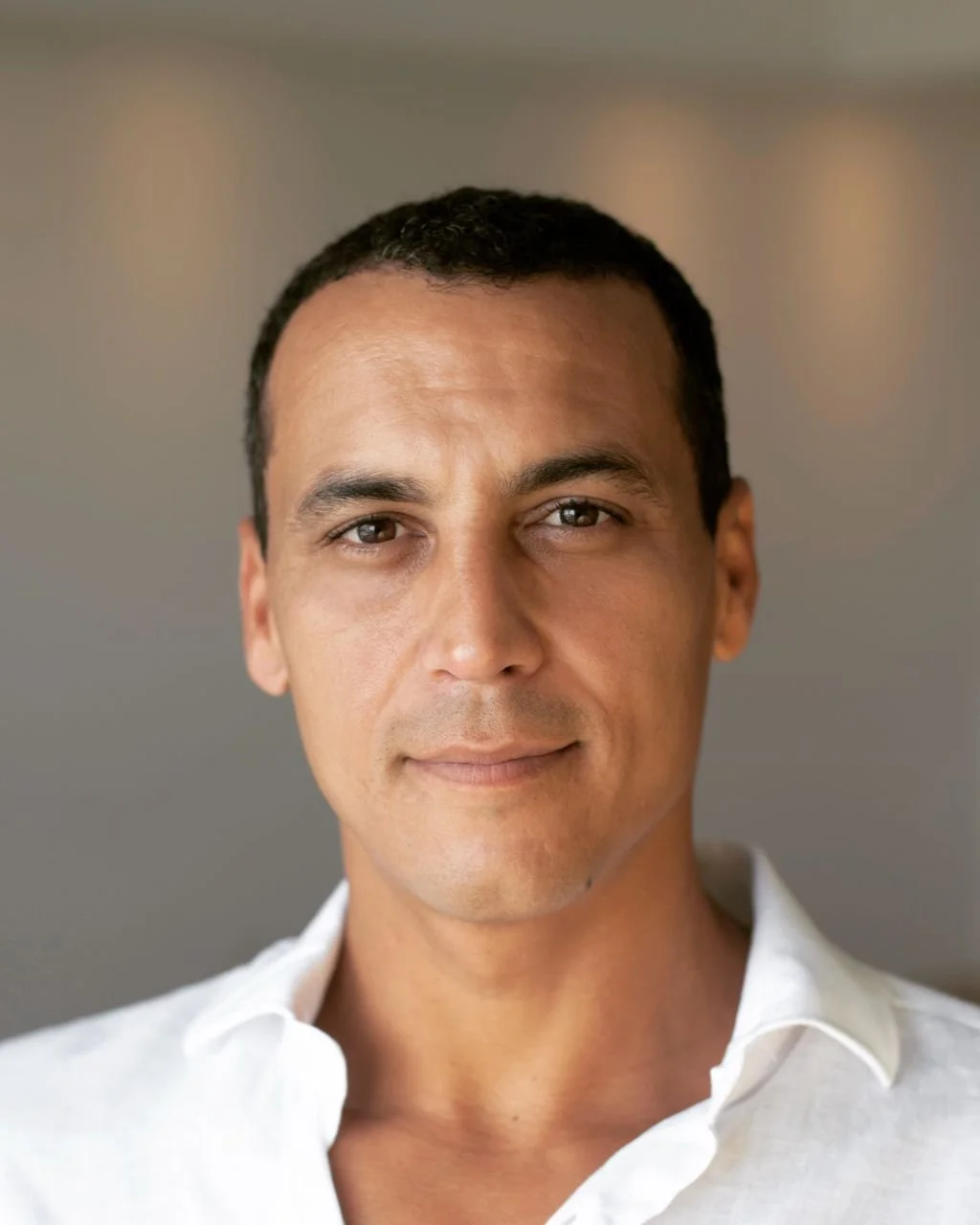 Close-up portrait of a man with short curly black hair, light brown skin, and brown eyes. He is wearing a white collared shirt and looking directly at the camera with a neutral expression.