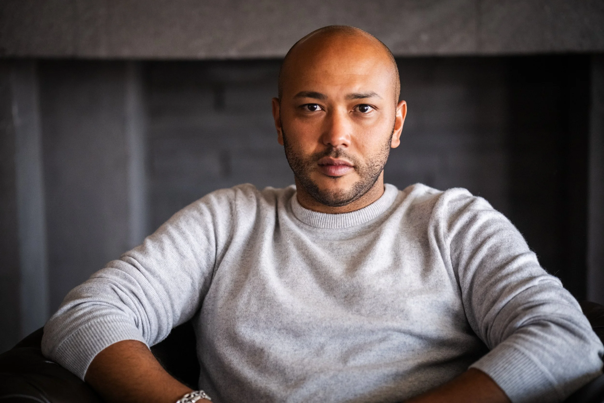 A man with a shaved head and beard, wearing a light gray sweater, sitting on a black leather chair inside a room with a dark gray background.