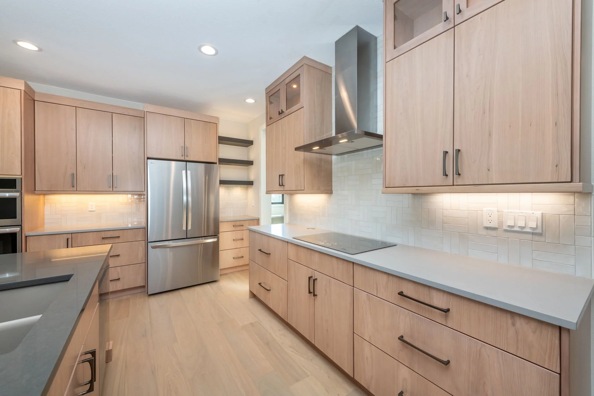Modern kitchen with light wood cabinets, stainless steel refrigerator, white countertops, light wood flooring, and a white subway tile backsplash.