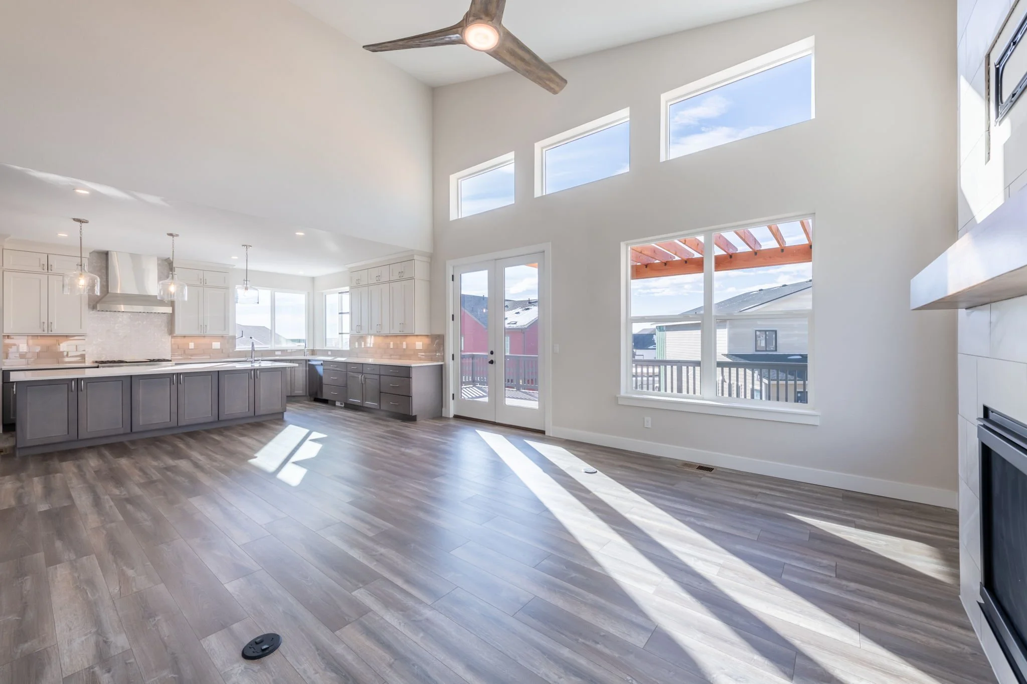 Bright, spacious kitchen with high vaulted ceiling, large windows, and sliding glass door leading to outdoor deck, featuring light-colored cabinetry, gray lower cabinets, stainless steel range hood, and wood-look flooring.