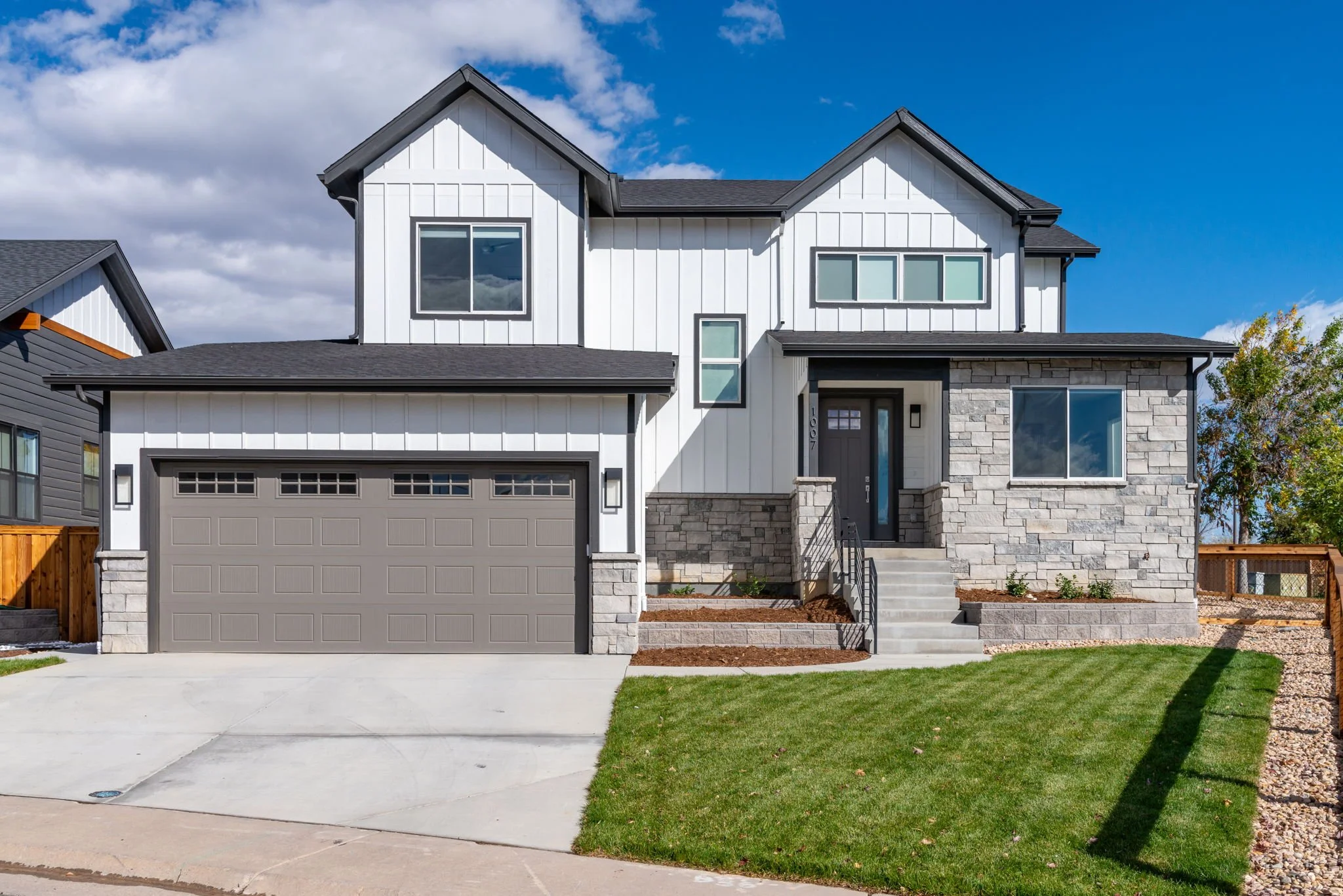 A modern two-story house with white siding, black roof, gray garage door, and stone accents. Front steps lead to a dark front door, with a green lawn and landscaped area. Marshall Fire rebuild