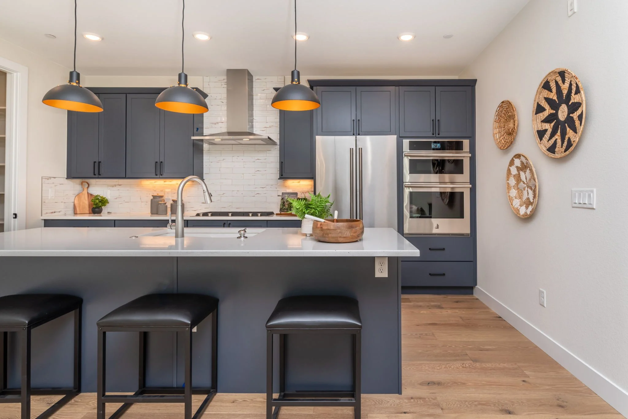 Modern kitchen with navy blue cabinets, white countertop, and stainless steel appliances, including double ovens. There are three black pendant lights hanging above the island with three black stools, a wooden bowl with green plants on the counter, a