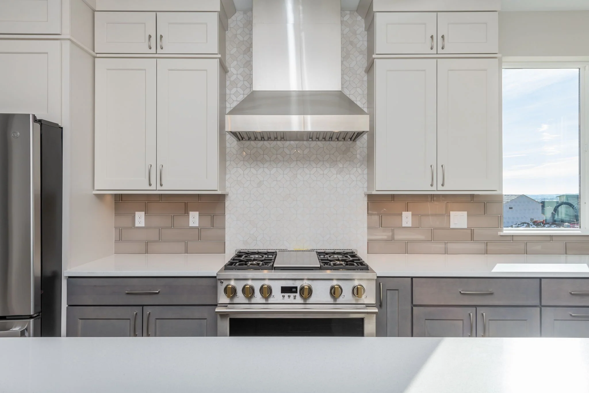 Modern kitchen with white upper cabinets, gray lower cabinets, a stainless steel stove with oven, a stainless steel range hood, tile backsplash, a window, and a view outside.