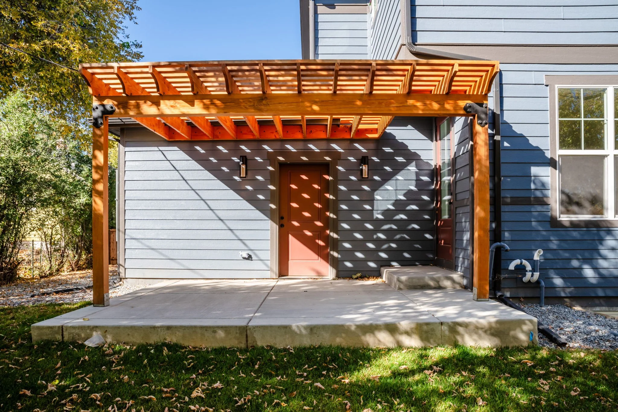 Backyard patio with a blue house, wooden pergola, door, window, and shadow patterns from the pergola on the wall.