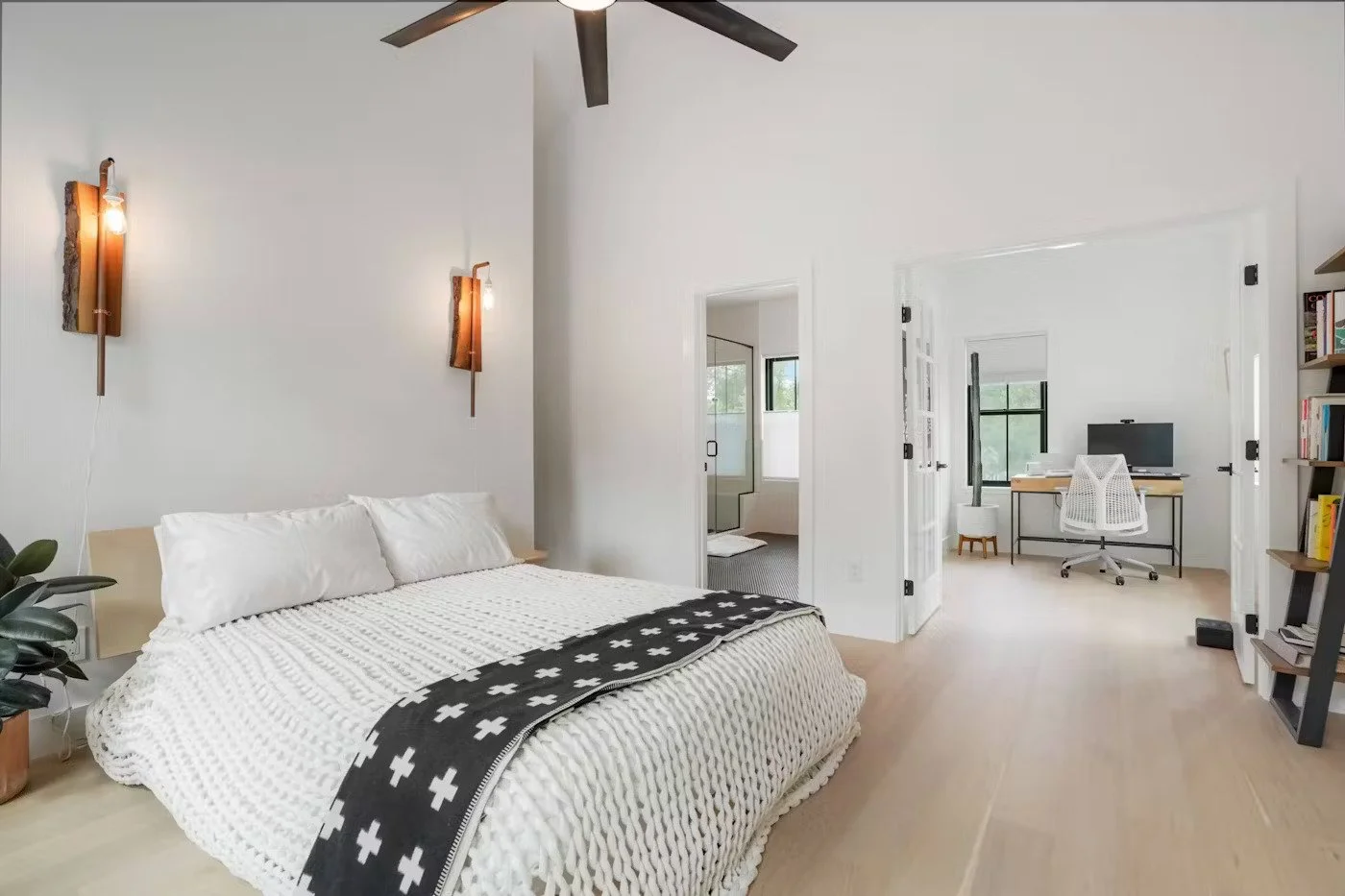 A bright, modern bedroom with a white bed, black and white blanket, wall-mounted lights, and a ceiling fan, with a doorway leading to a home office area and a bathroom.
