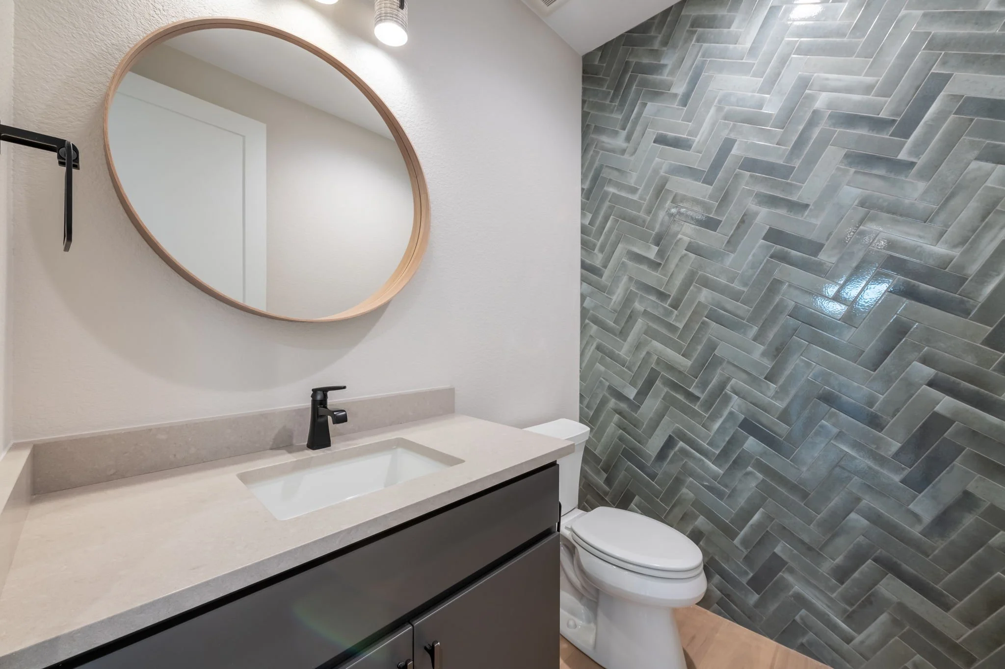 A modern bathroom with a white sink and black faucet on a beige countertop, a large oval mirror with a wooden frame, white walls, a partial view of a wooden floor, and a wall with gray herringbone tile behind the toilet.