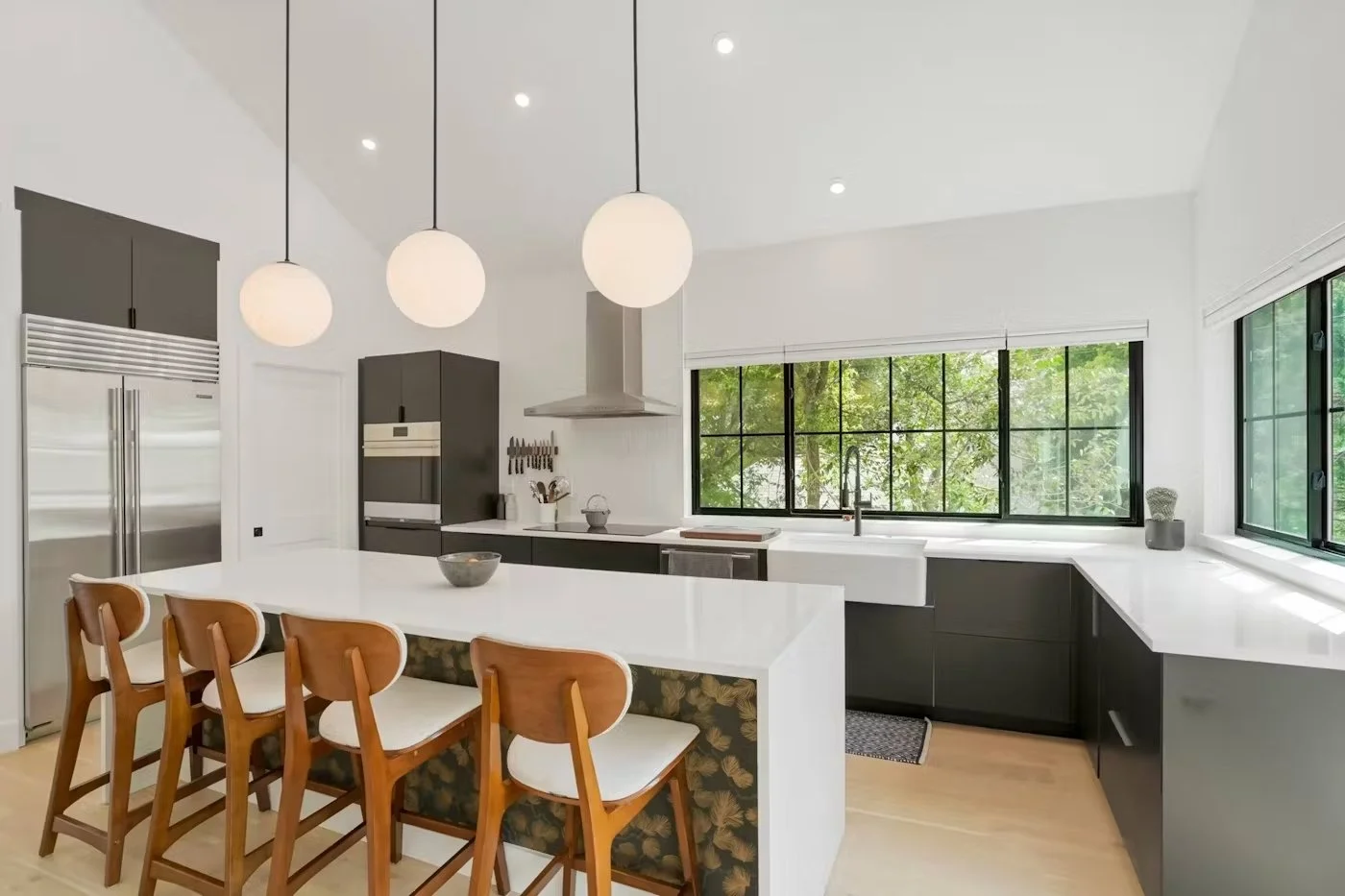 Modern kitchen with white countertops, black cabinets, large windows with greenery outside, island with four wooden chairs, stainless steel refrigerator, and pendant lights.