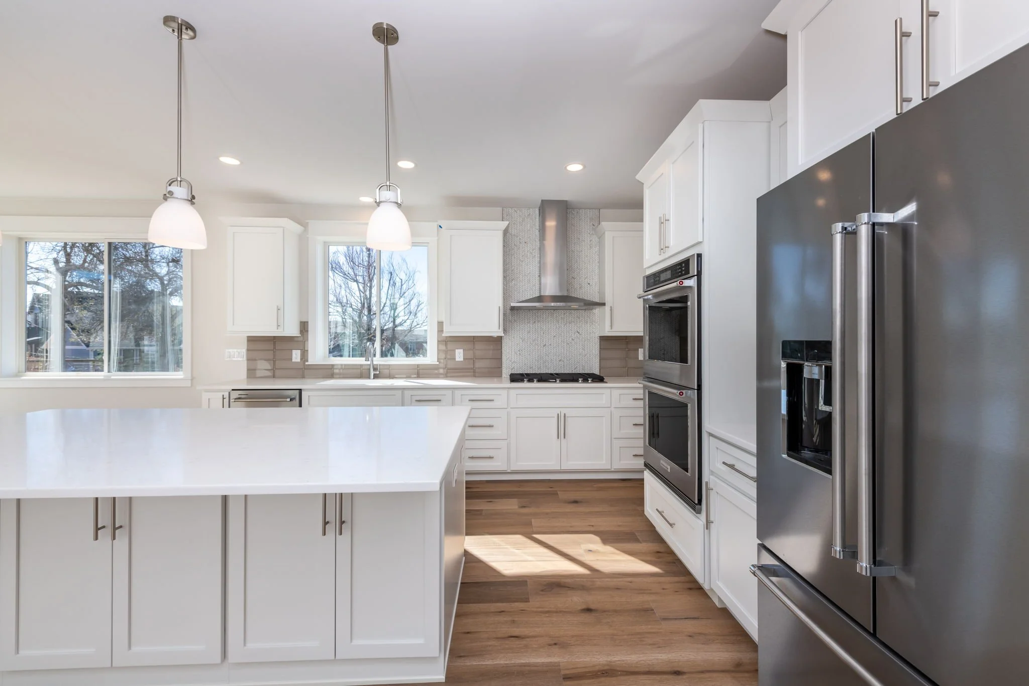 Modern kitchen with white cabinets, stainless steel appliances, double wall ovens, a large refrigerator, a kitchen island, wood flooring, and pendant lighting.