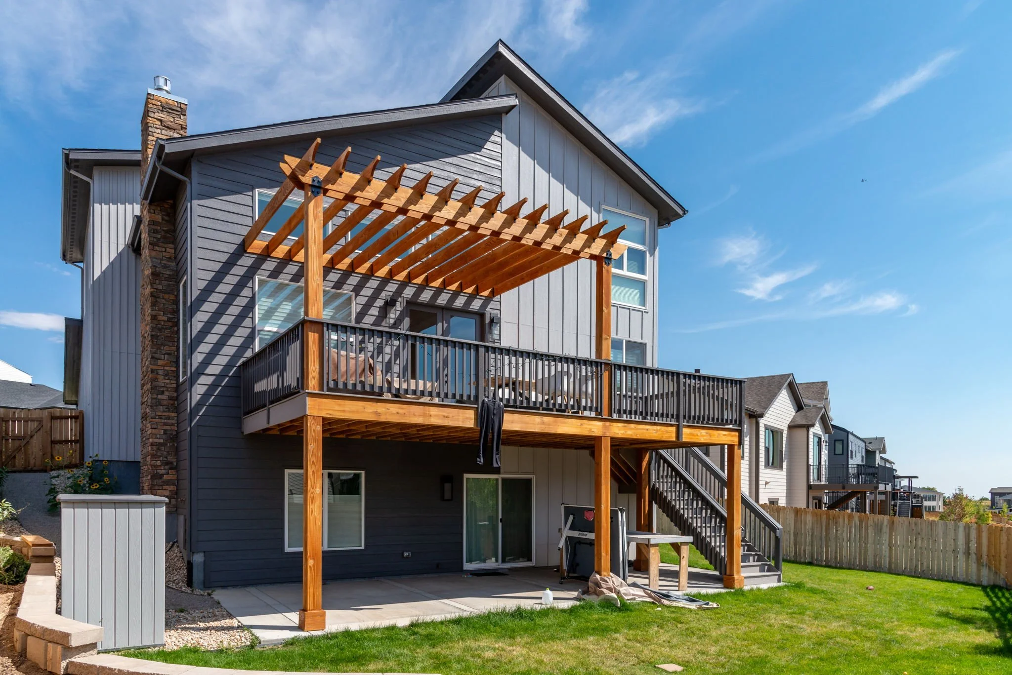Rear view of a modern multi-story house with a spacious backyard, wooden deck, and pergola under construction, sunny day with blue sky.