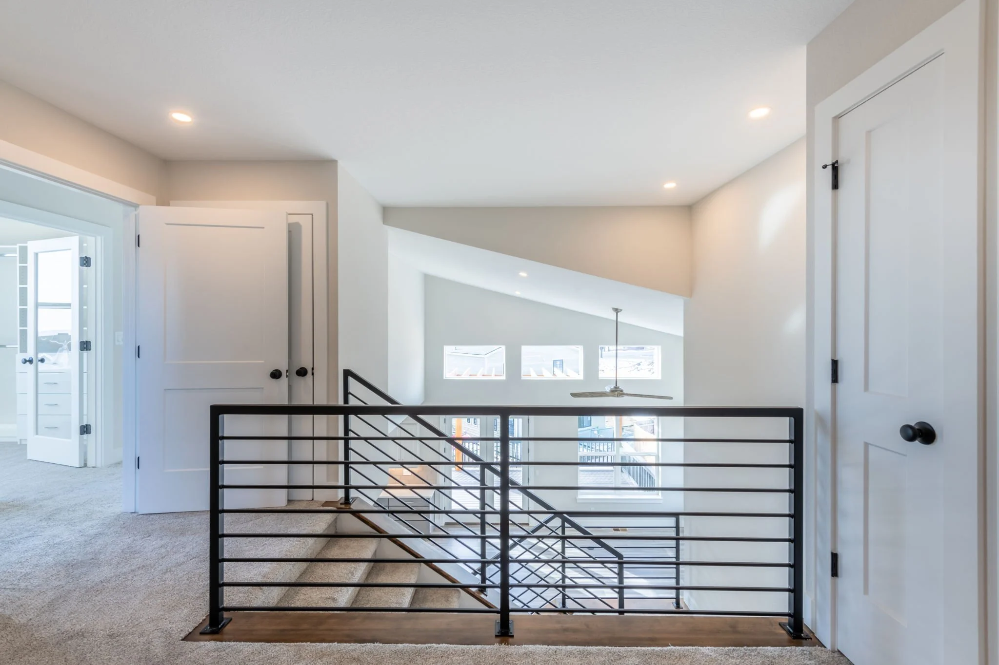 Interior view of a modern home upper floor with open railing, carpeted floor, closed white doors, and a partial view of a lower level staircase with large windows letting in natural light.