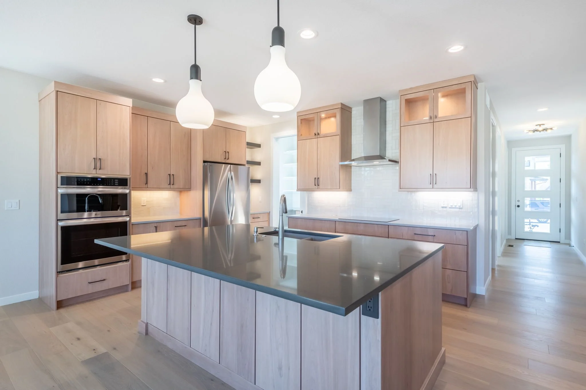 Modern kitchen with light wood cabinetry, stainless steel appliances, black countertop island, and pendant lights.