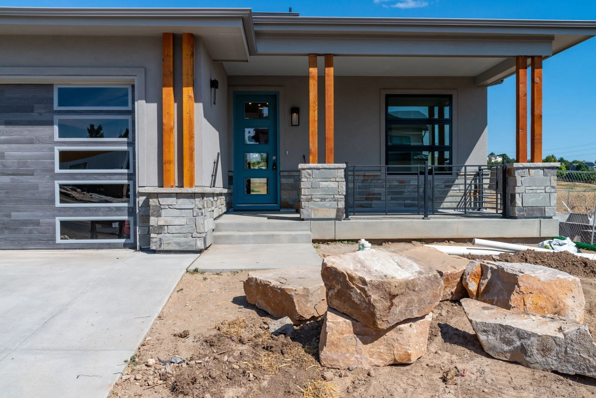 A modern house under construction with a front porch, concrete steps, a blue front door, stone accents, and large window openings. The yard is unfinished with rocks, dirt, and construction materials.