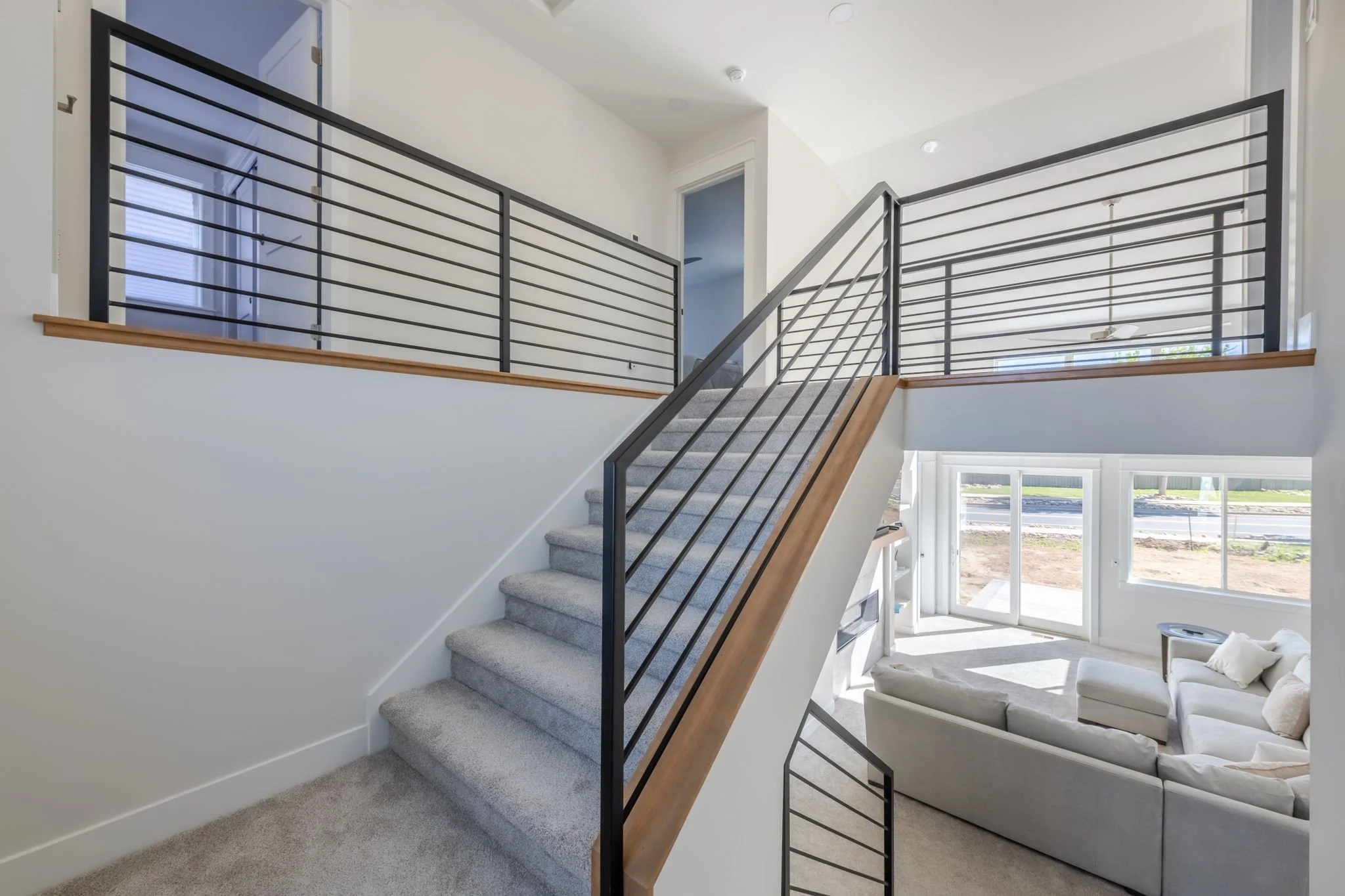 Interior view of a modern two-story living space with a staircase, gray carpet, white walls, black metal railings, large windows, and a seating area with a sectional sofa.