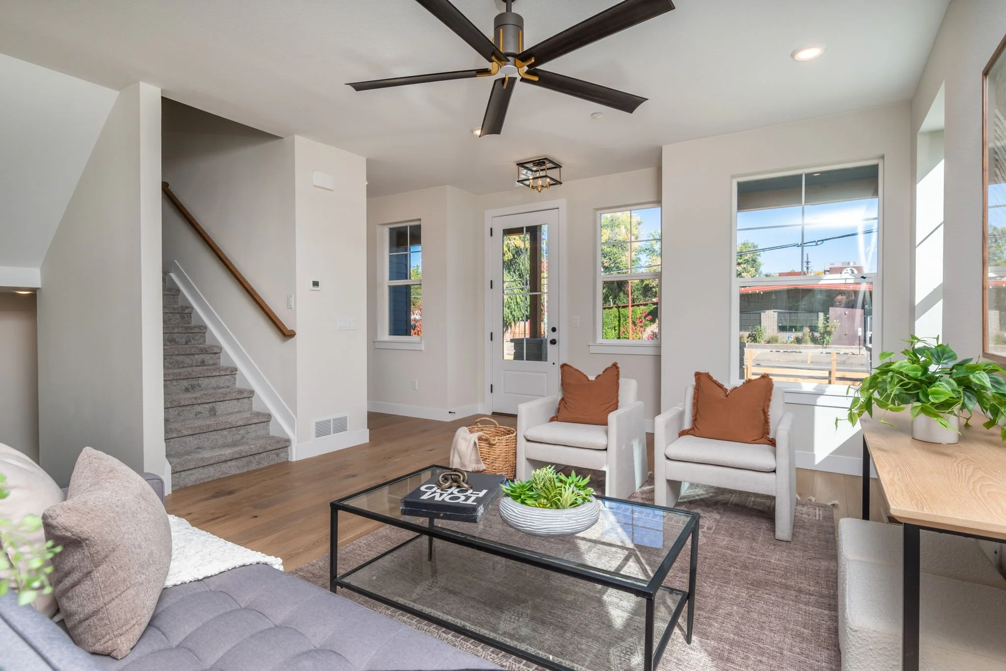 Living room with white walls, large windows, a ceiling fan, a glass coffee table, a gray sofa with beige pillows, two white armchairs with orange pillows, a wooden console with a potted plant, and a staircase with carpeted steps.