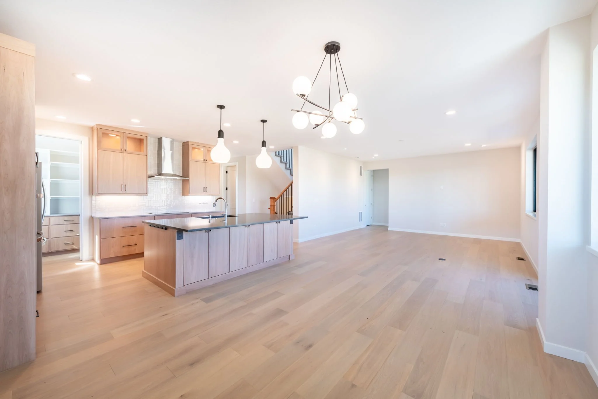 Open-concept living space with a modern kitchen and hardwood floors. The kitchen has light wood cabinets, a kitchen island with a sink, and pendant lighting. The living area is spacious with white walls and large windows.