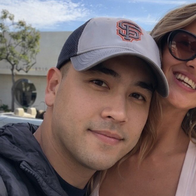 A young man wearing a baseball cap and a hoodie, sitting indoors with a blurred background of ceiling lights.