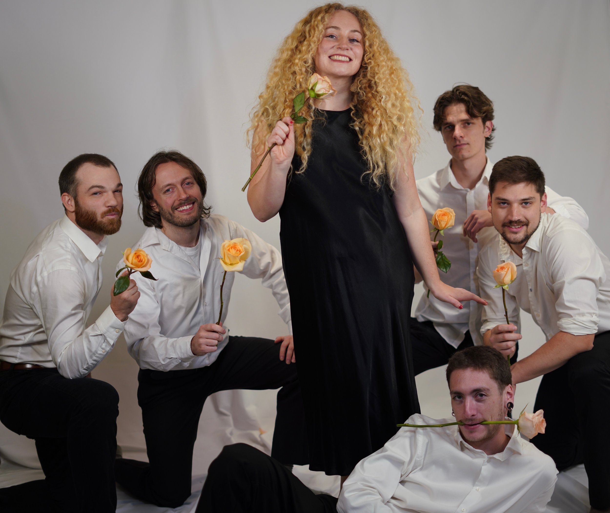 Group of six people, five men and one woman, with roses, posing against a plain backdrop. The woman in the center has curly blonde hair and is wearing a black dress, smiling. The men are surrounding her, dressed in white shirts, some kneeling, some sitting or standing, holding or with roses, with playful expressions.