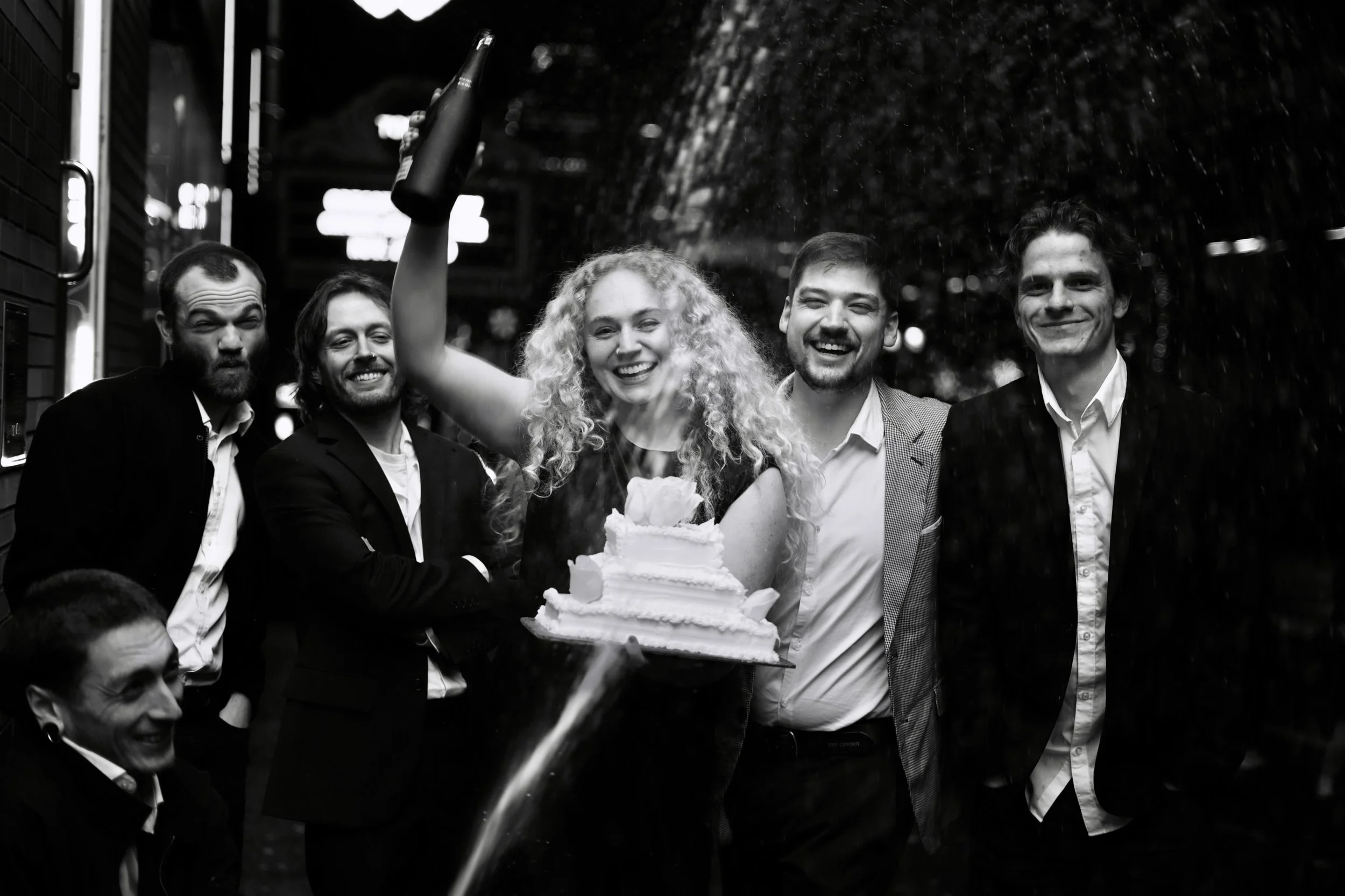 A group of six people, five men and one woman, celebrating at night. The woman, in the center, has curly hair and is holding a cake with a slice on top. She is smiling as she receives a bottle of champagne. The group appears joyful and are dressed in formal and semi-formal attire.