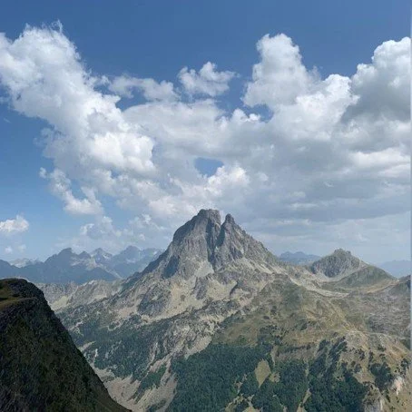 Mountain landscape with a prominent peak, green slopes, and fluffy clouds in the sky.