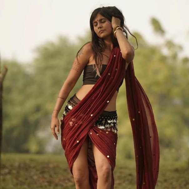 A woman in traditional Indian attire outdoors, wearing a black crop top and a red saree with embroidery, standing in a natural setting with trees in the background.