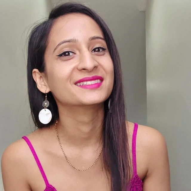 A woman with long dark hair wearing pink lipstick, a pink tank top, large earrings, and a delicate gold necklace, smiling at the camera.