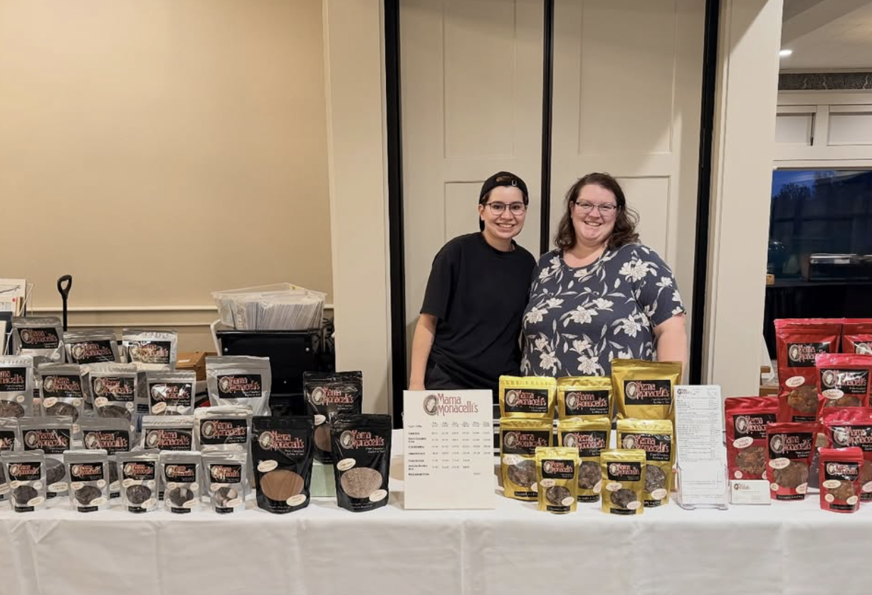 Two women standing behind a table displaying packaged baked goods, including cookies, at an indoor event. One woman is wearing a black shirt and backwards cap, the other a floral shirt. A sign with the name 'Mama Monacellis' is on the table.