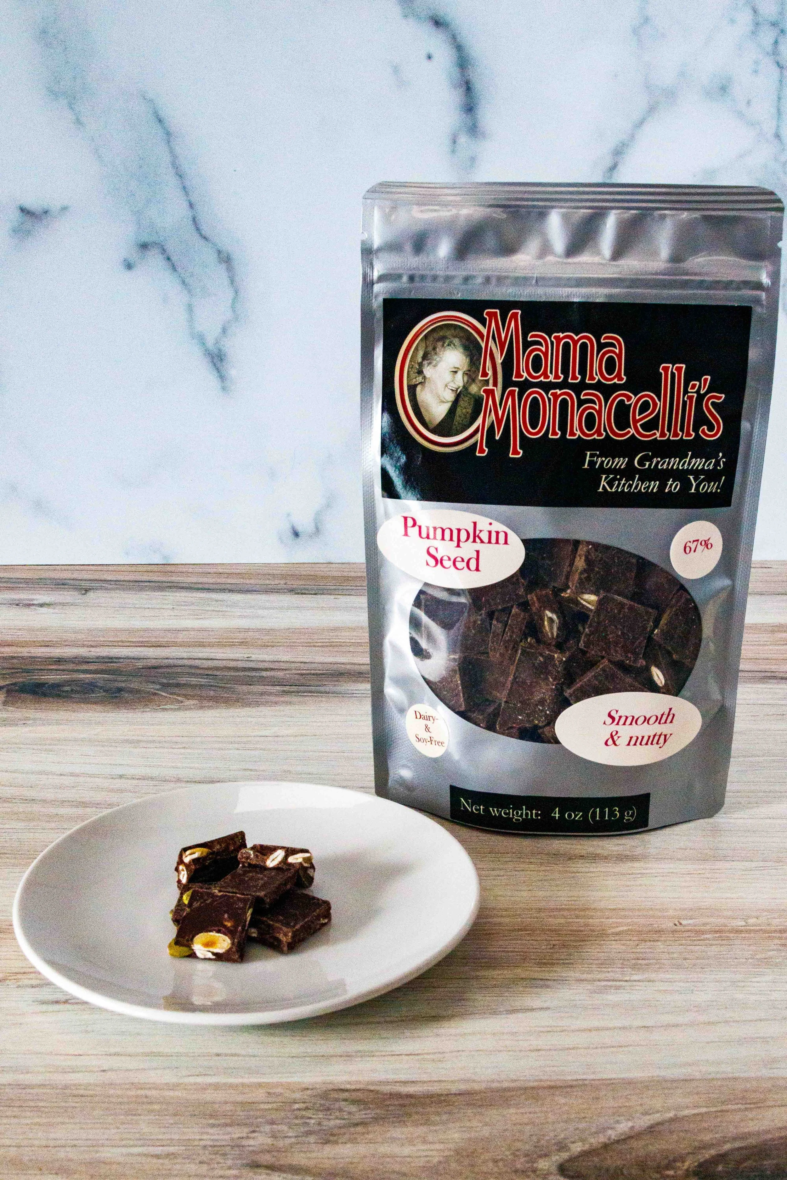 A bag of Mama Monacelli's Pumpkin Seed chocolate pieces on a wooden surface with some chocolate pieces on a small white plate in front.