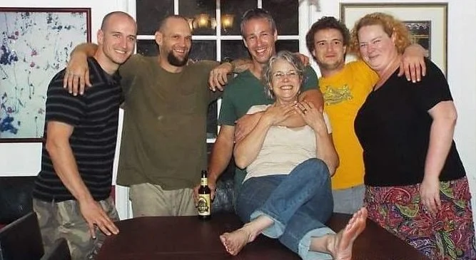 Six people gathered around a table, smiling. One woman is sitting on the table with her arms around a man, and the others stand behind her with arms around each other. The group appears happy and relaxed in a casual setting.