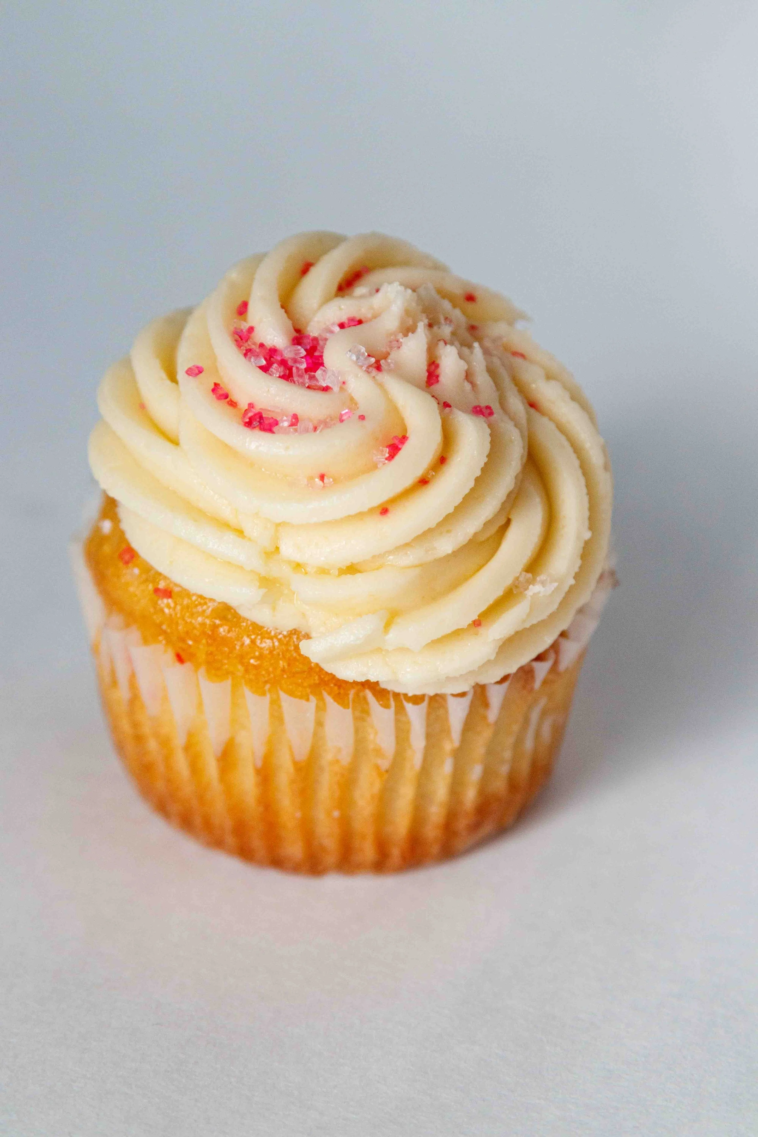 A vanilla cupcake with white frosting swirled on top, decorated with pink and red sprinkles.