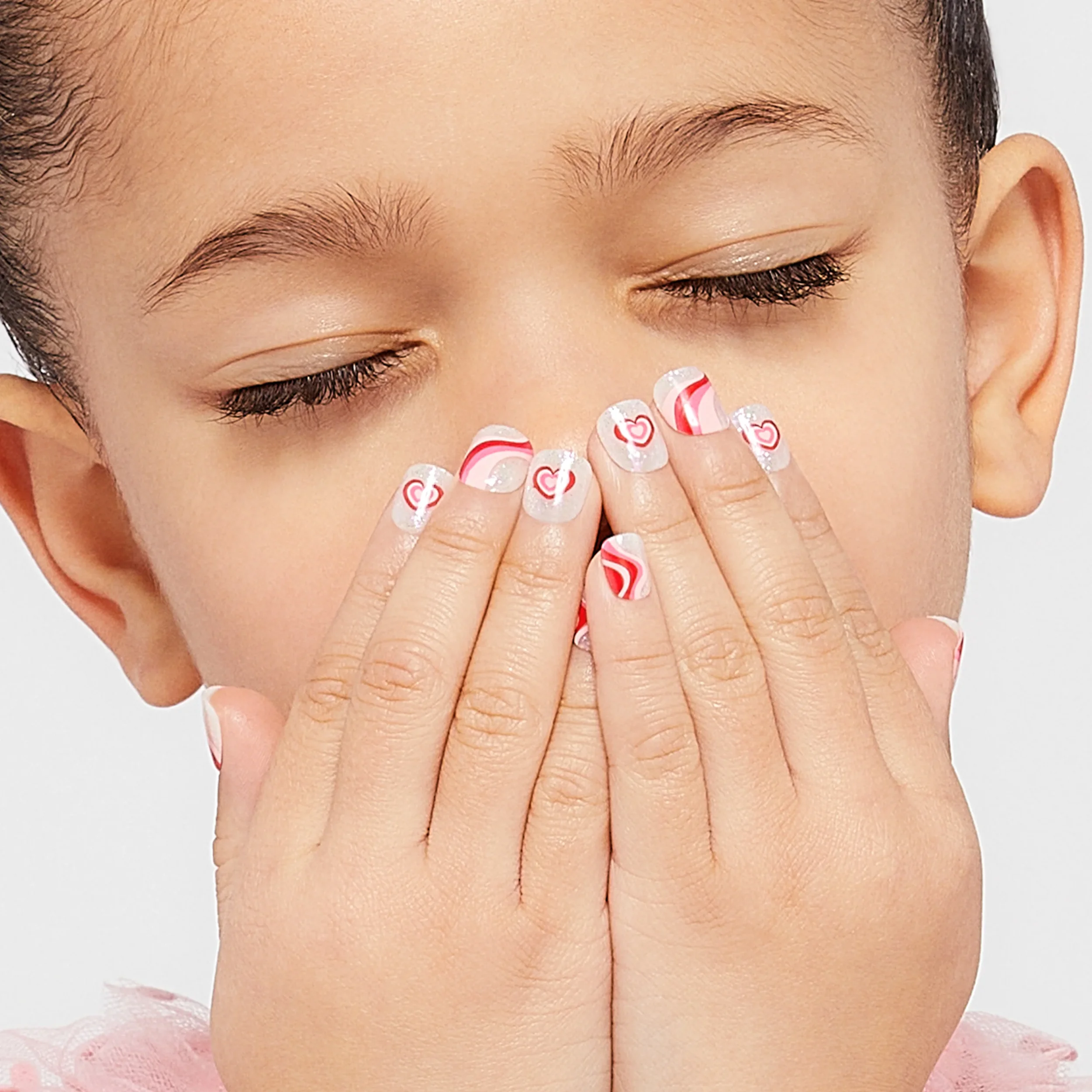 A young girl with closed eyes, covering her nose and mouth with her hands. She has decorated nails with pink and red swirled and heart patterns.