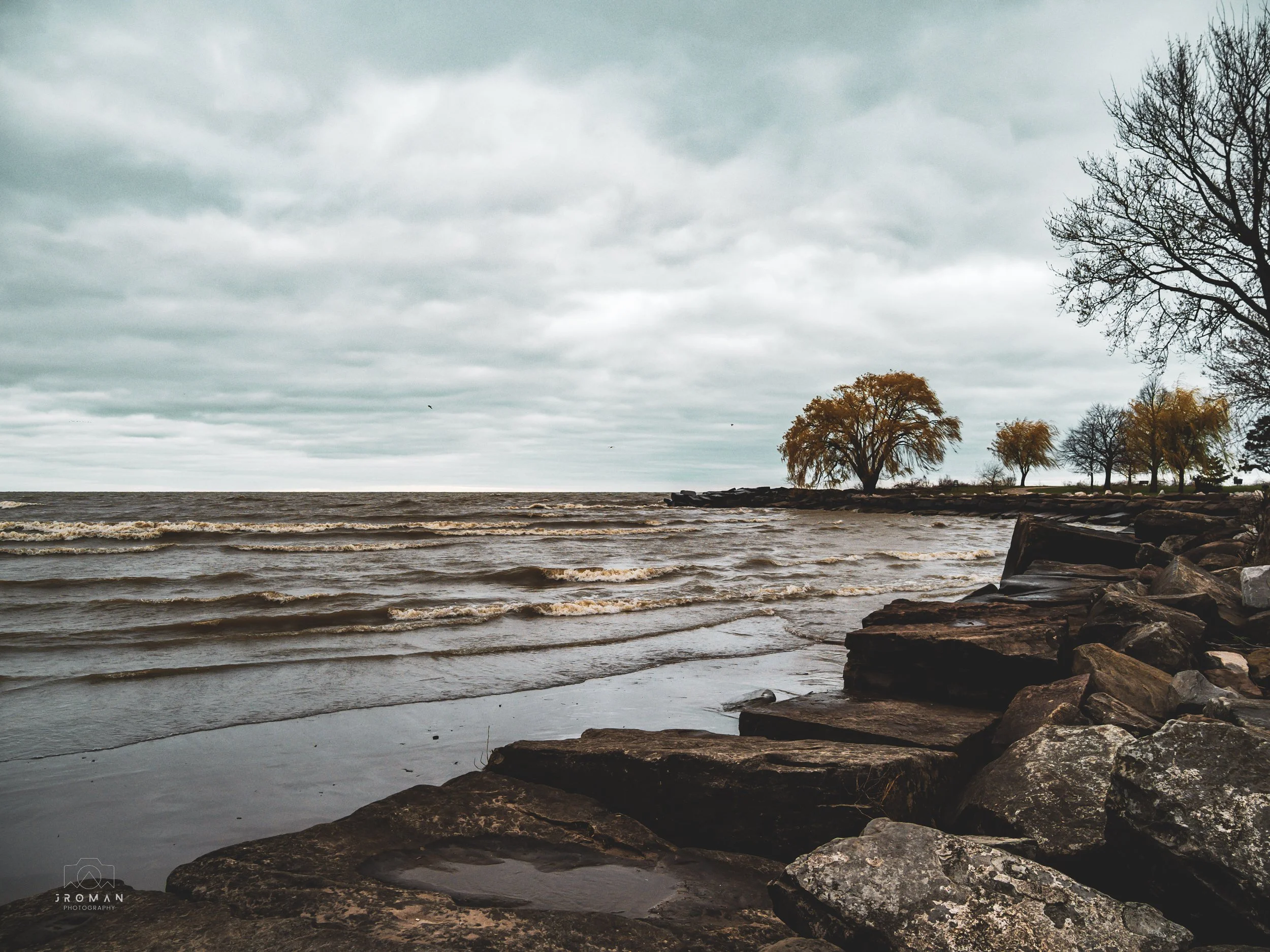 Overcast day at a lakeshore with small waves, rocks along the shore, and trees with yellow and bare branches in the distance.