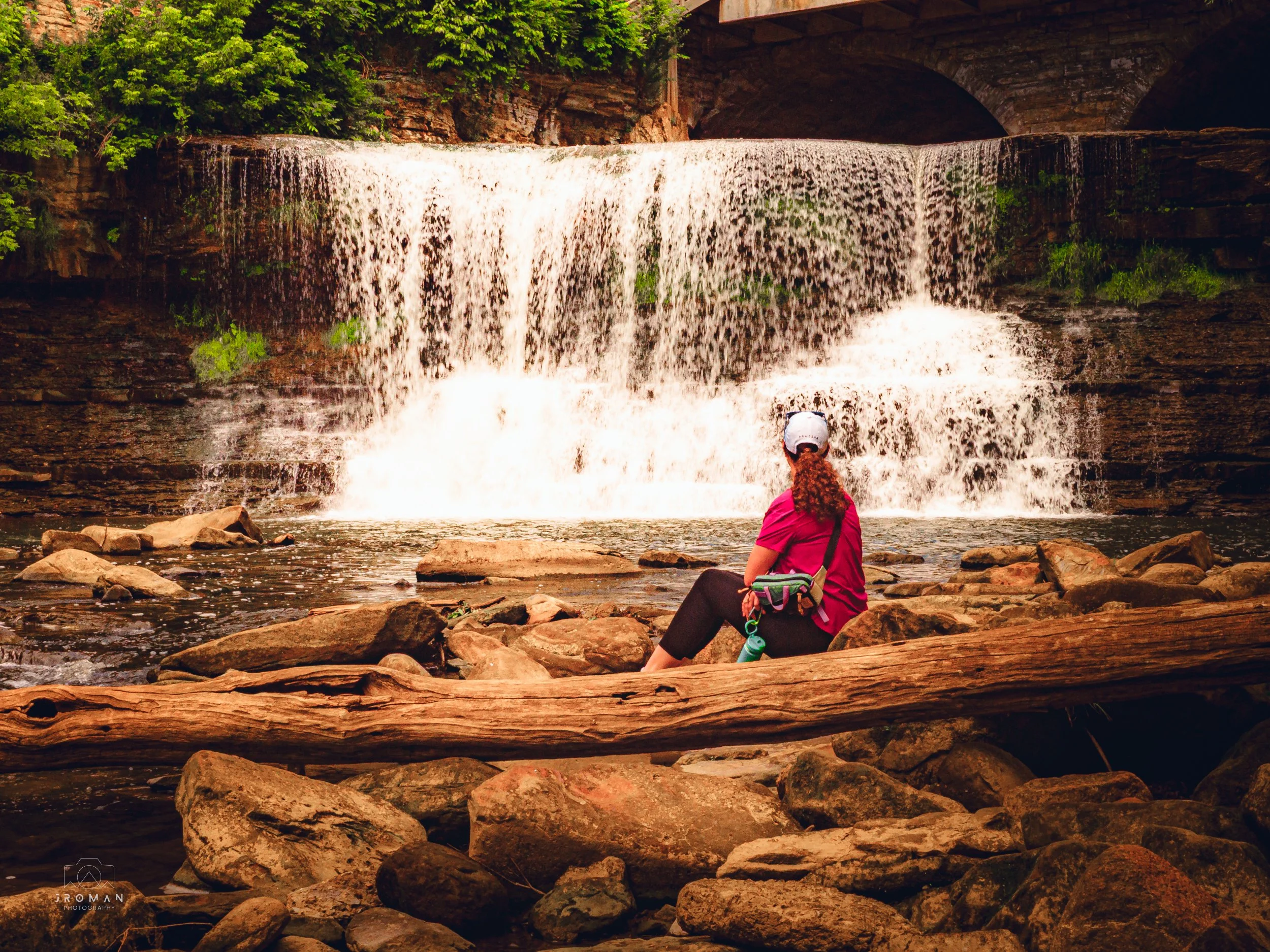 A woman with curly red hair wearing a white cap, red shirt, and black pants sits on rocks near a small waterfall surrounded by trees.