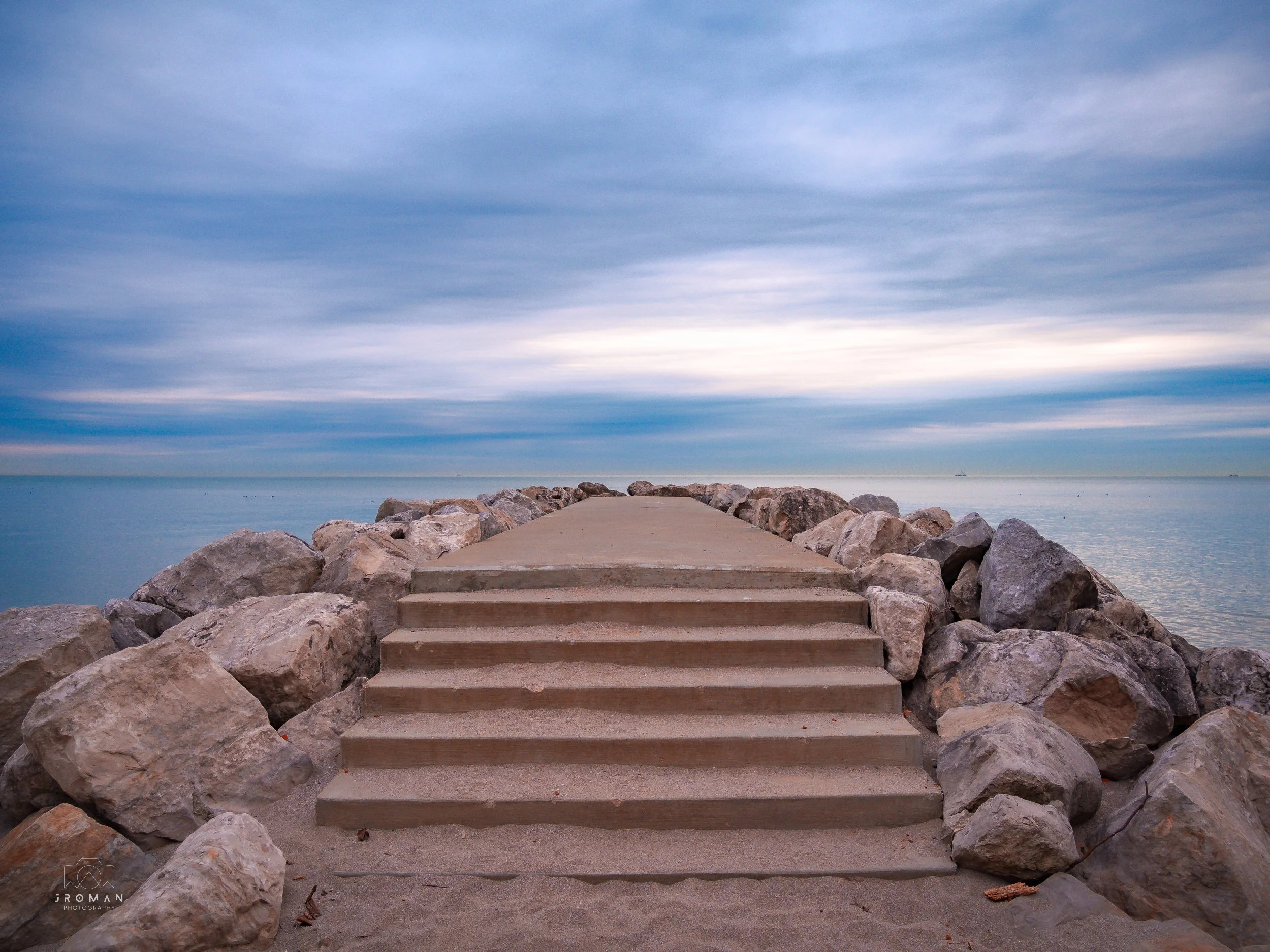 Concrete steps leading to a rocky pier extending into calm water under a cloudy sky.