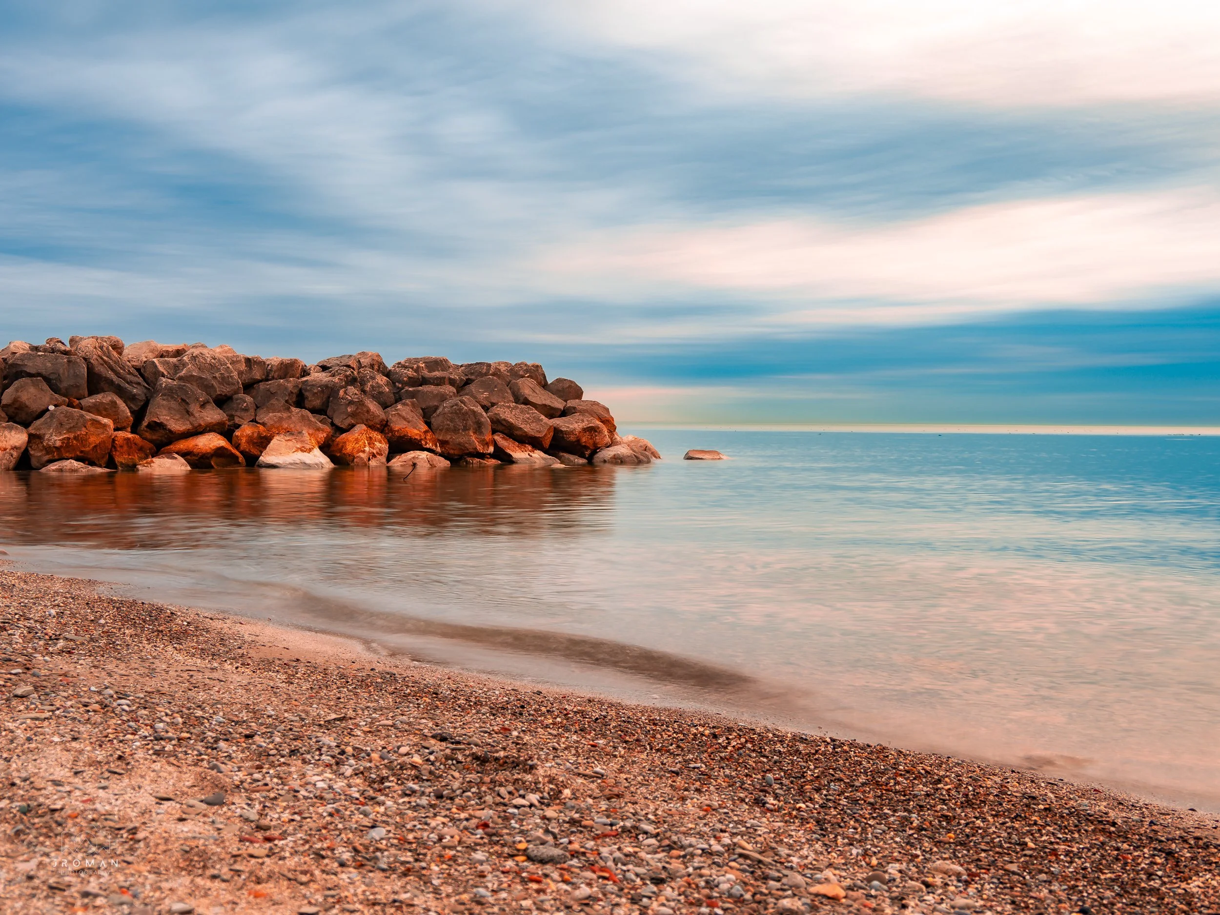 A beach with pebbles and rocks, calm water, and a rock jetty, under a cloudy sky