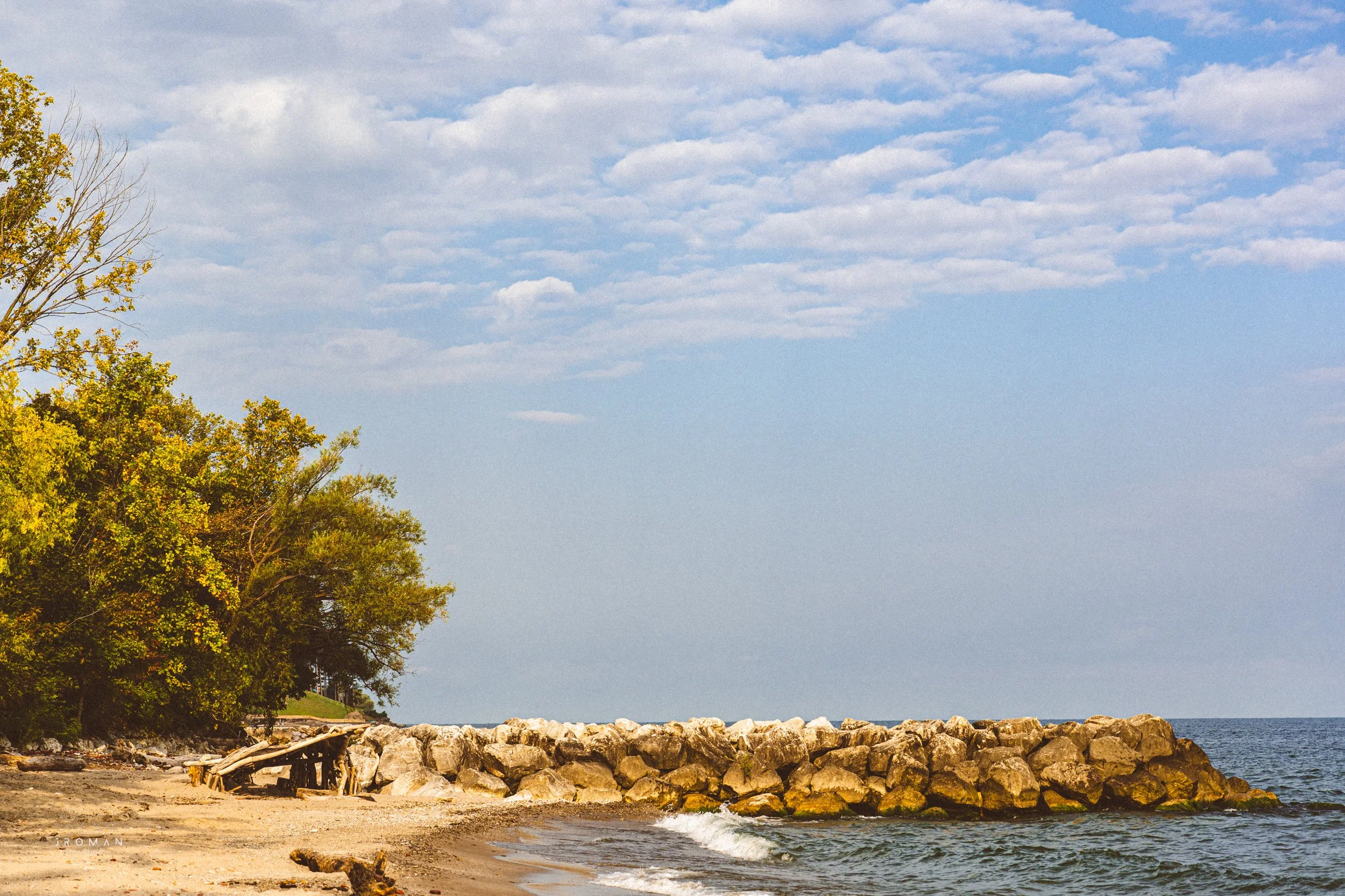 A peaceful beach scene with a sandy shoreline, large rocks forming a breakwater, trees with green and yellow leaves on the left, and the ocean stretching towards the horizon on the right, under a partly cloudy sky.