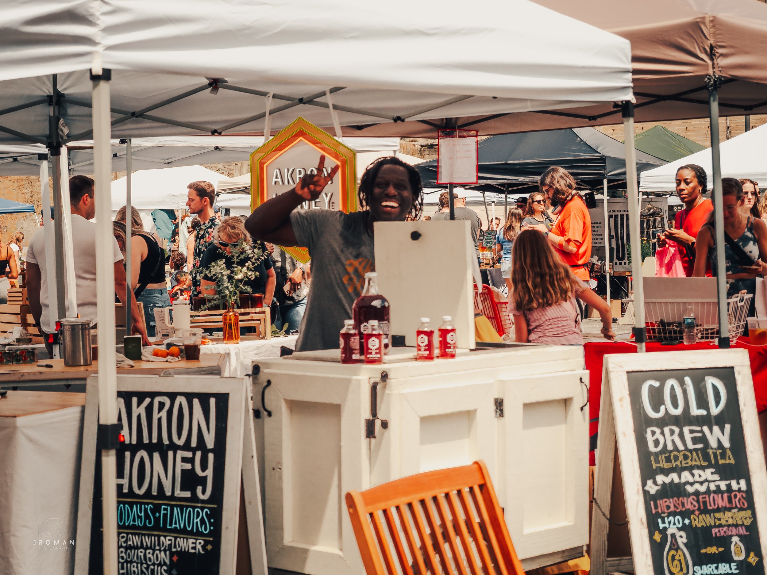 A lively outdoor market with various vendor tents. A smiling person with dreadlocks is behind a stand selling Akron Honey, with bottles of honey on display. Chalkboard signs advertise cold brew herbal tea and Akron Honey with various flavors. Many pe