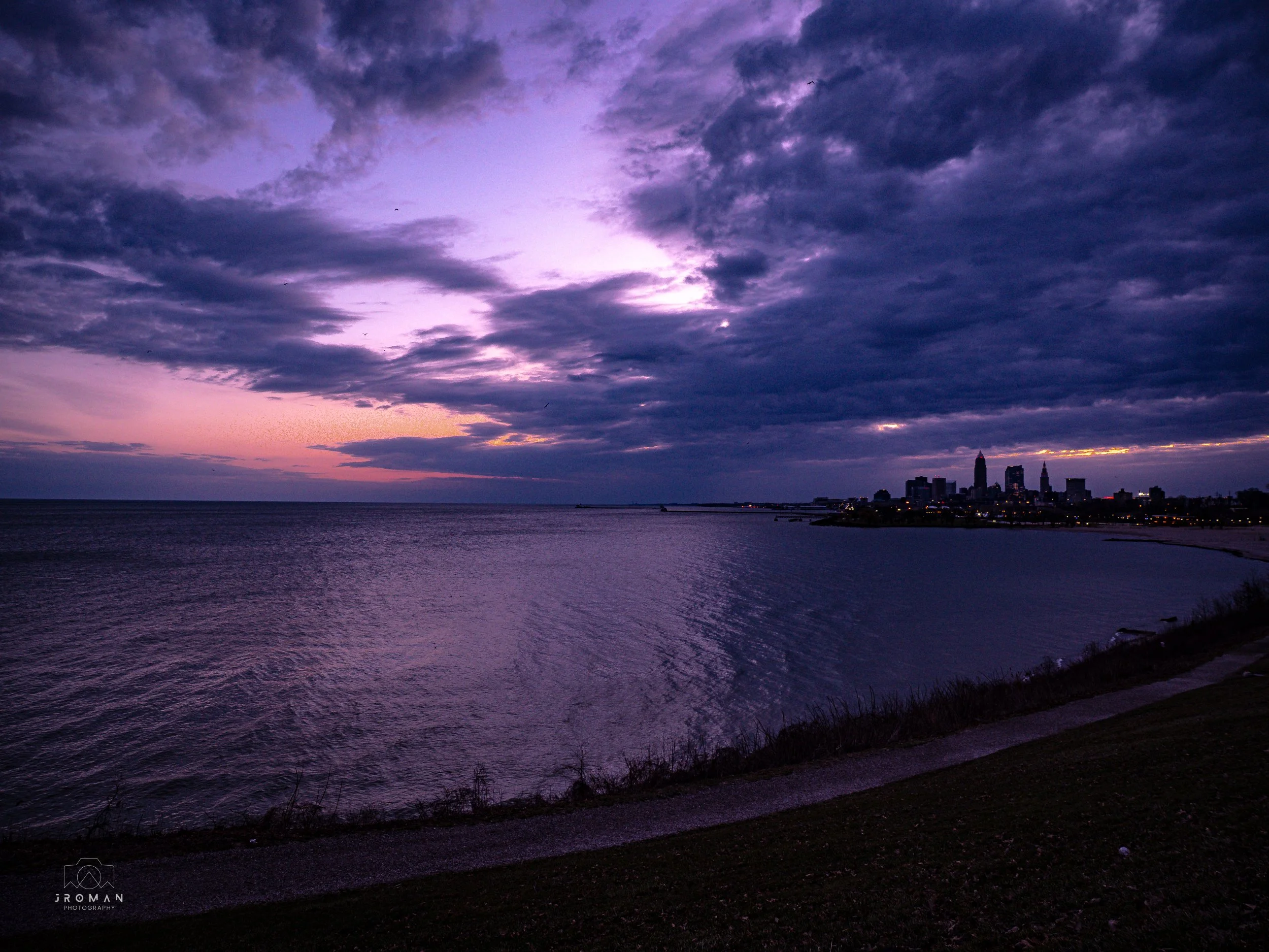 A purple and blue dusk sky over a calm body of water with a distant city skyline visible on the horizon.