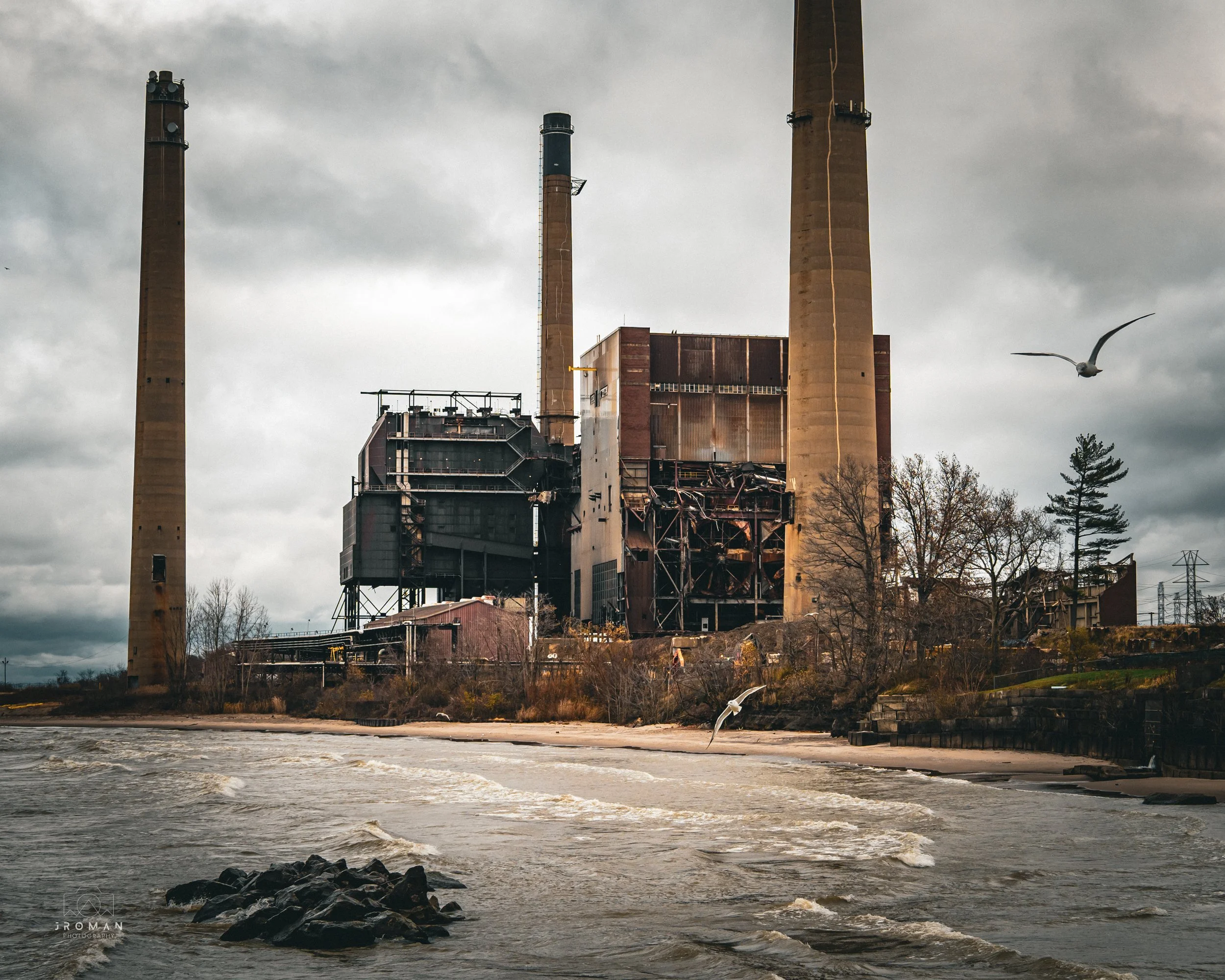 An abandoned industrial power plant with four tall, rusty smokestacks, located near a body of water. Overcast sky, leafless trees, and seagulls flying.