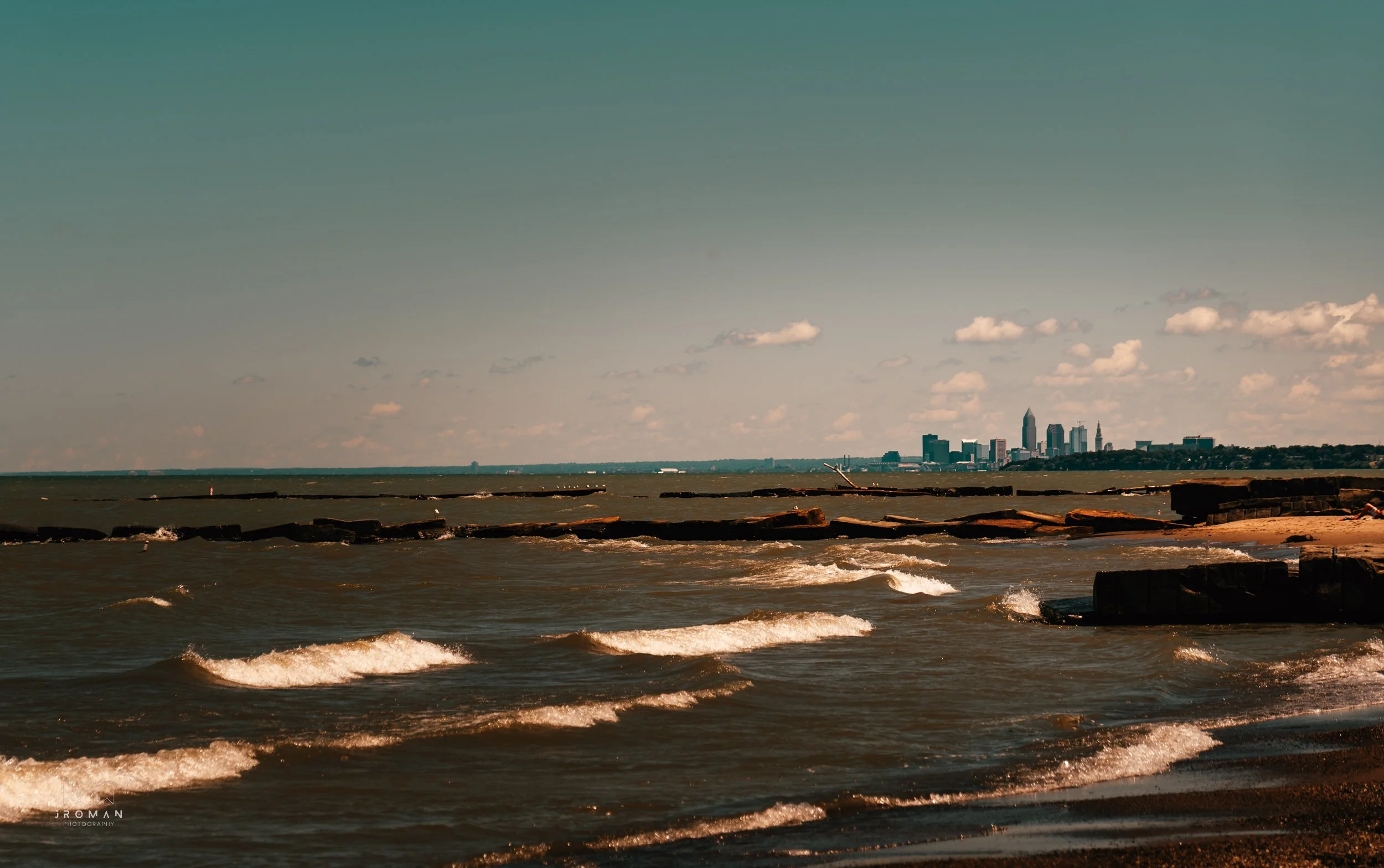 City skyline viewed from a beach with gentle waves, rocks, and a partly cloudy sky.
