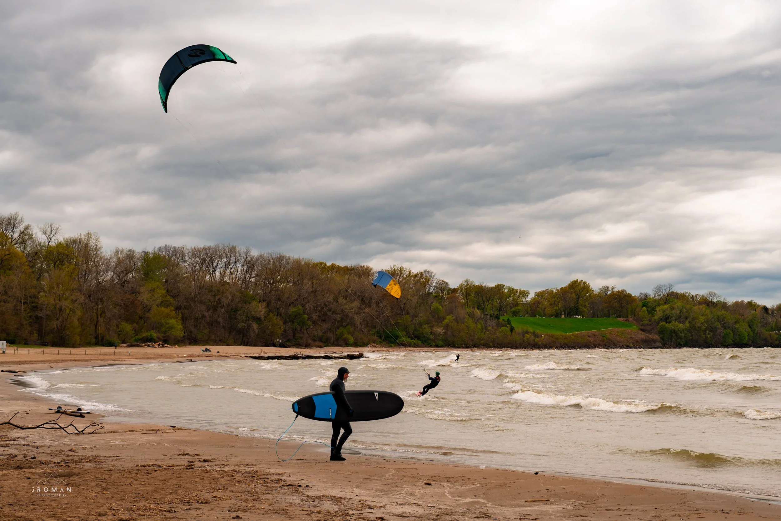 People kiteboarding on a beach with cloudy skies and trees in the background.