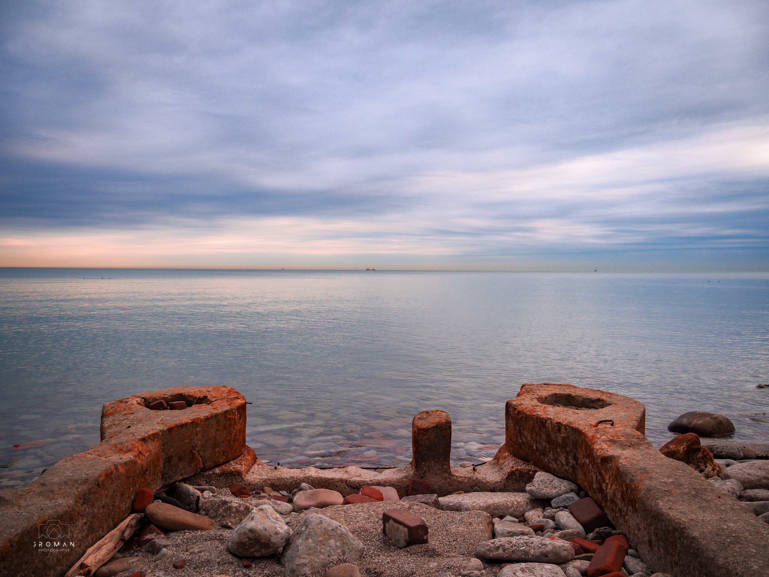 View of the water and sky with some rocks and rusted metal structures on the shore in the foreground.