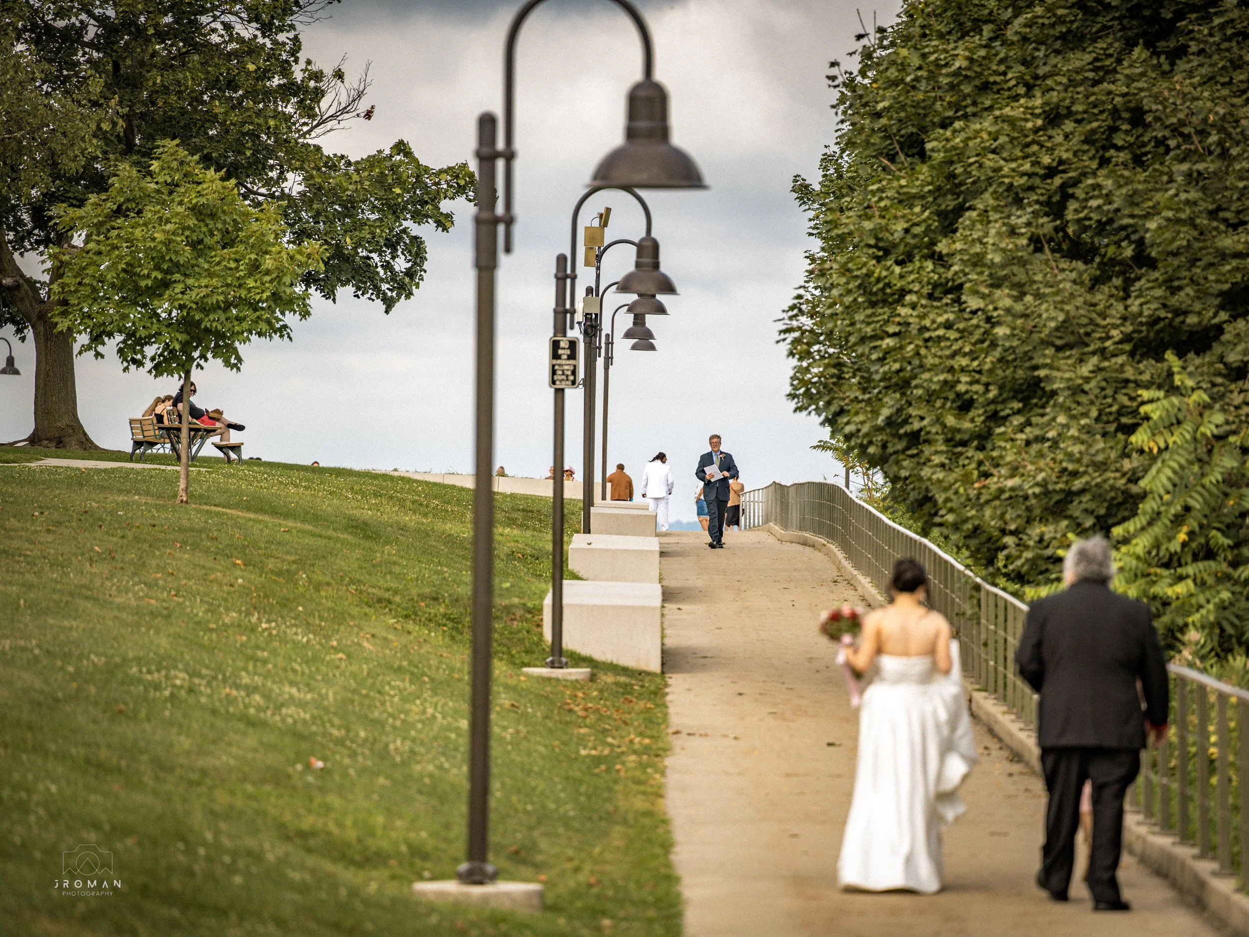 Couple in wedding attire walking along a paved pathway in a park, holding hands; woman holds a bouquet of flowers, man wears a suit; park has green grass, trees, lampposts, and some benches, with a cloudy sky.