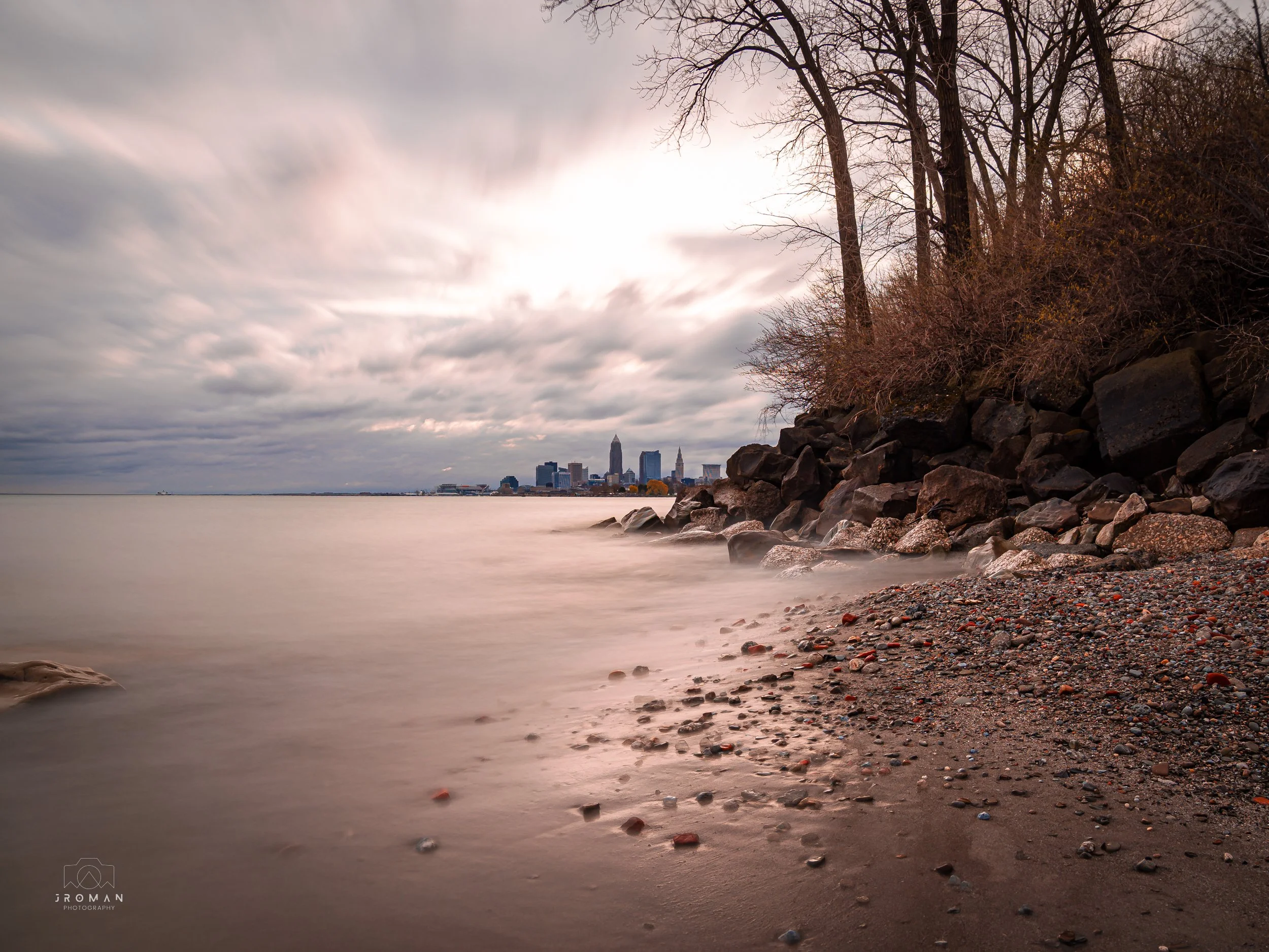 A rocky beach with a city skyline and cloudy sky in the distance.
